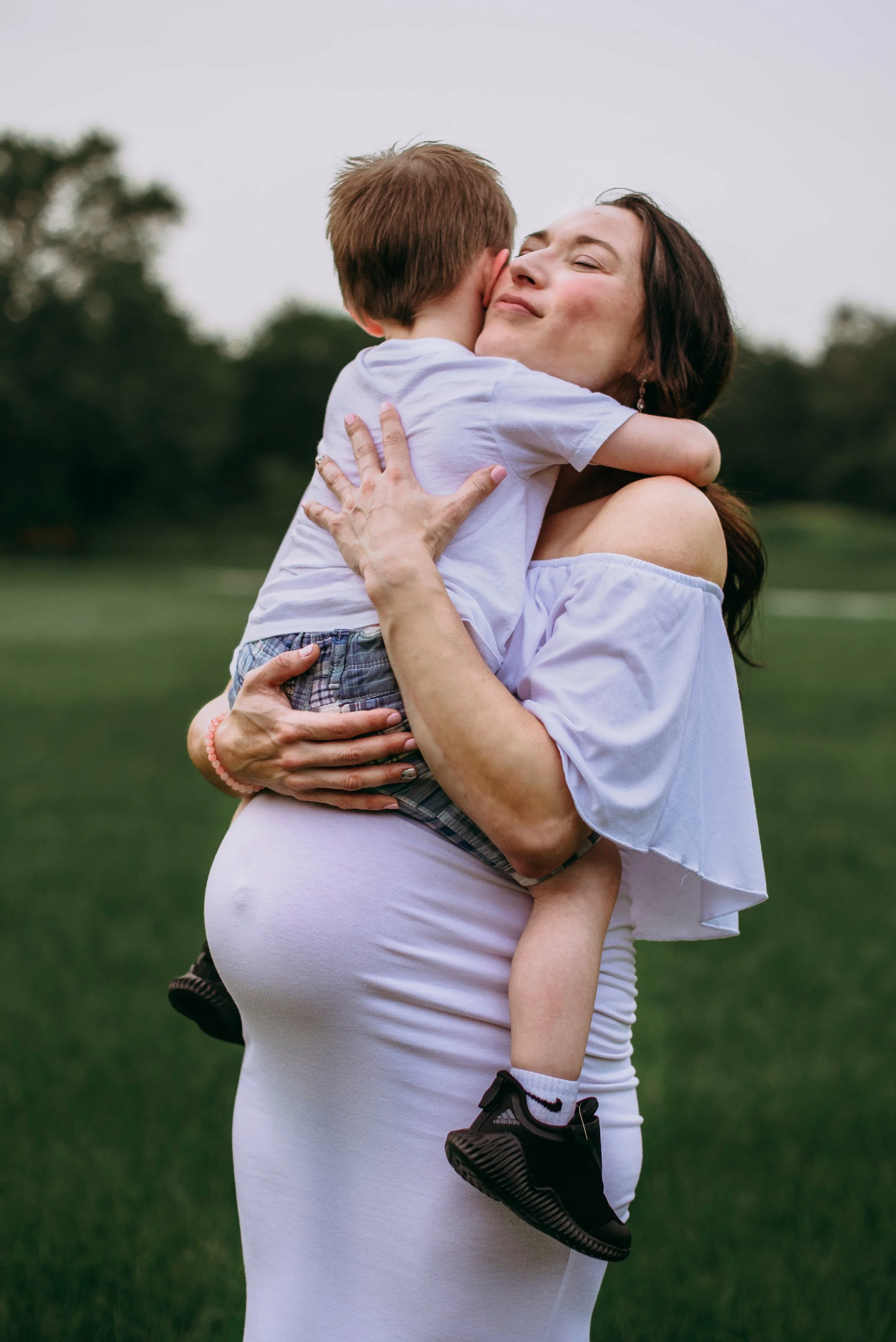 A woman with dark brown hair wearing a white off-shoulder top and white pants is hugging a young boy with brown hair, a white t-shirt, plaid shorts, and black sneakers with white socks. They are outdoors on a grassy field with trees in the background