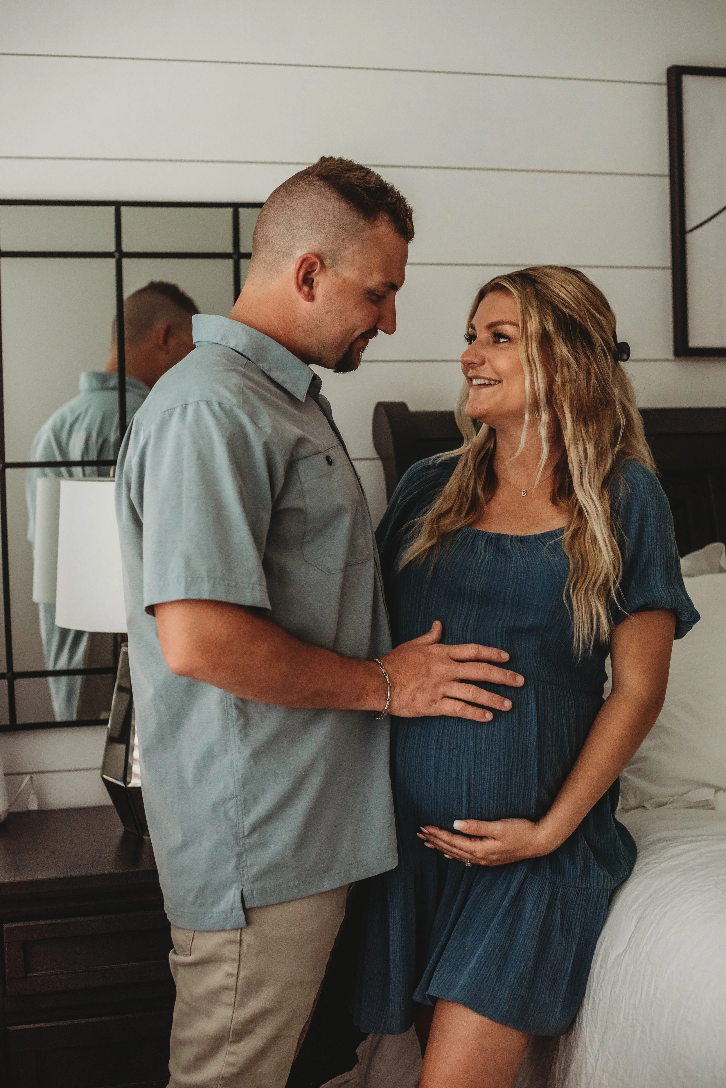 A couple, with the woman visibly pregnant, smiling and looking at each other in a bedroom. The woman is sitting on the bed, wearing a blue dress, and the man is standing in front of her, touching her belly. A mirror reflects part of the man's face.
