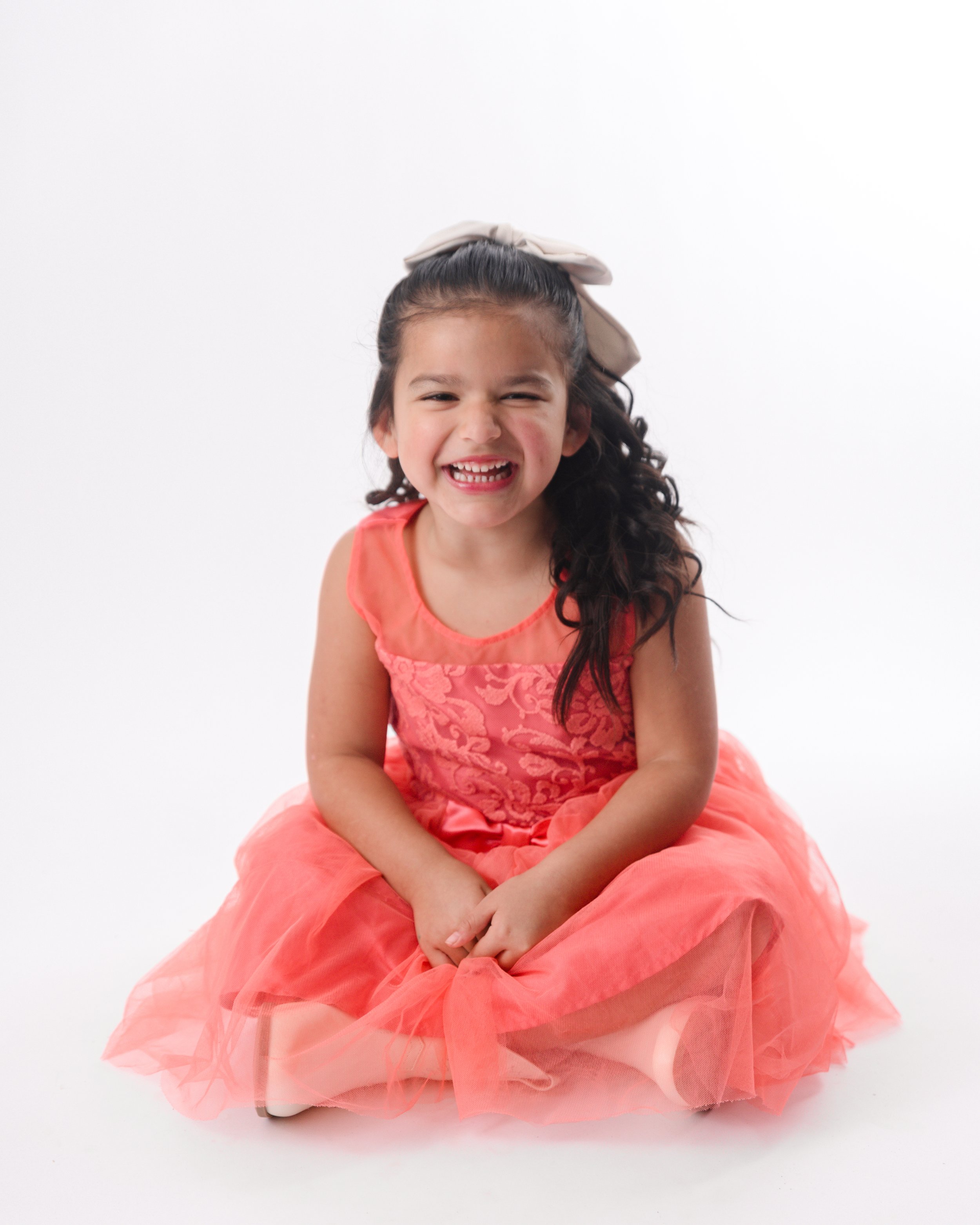 A young girl with long curly dark hair, wearing a coral pink dress and a beige bow in her hair, sitting on the floor and laughing.