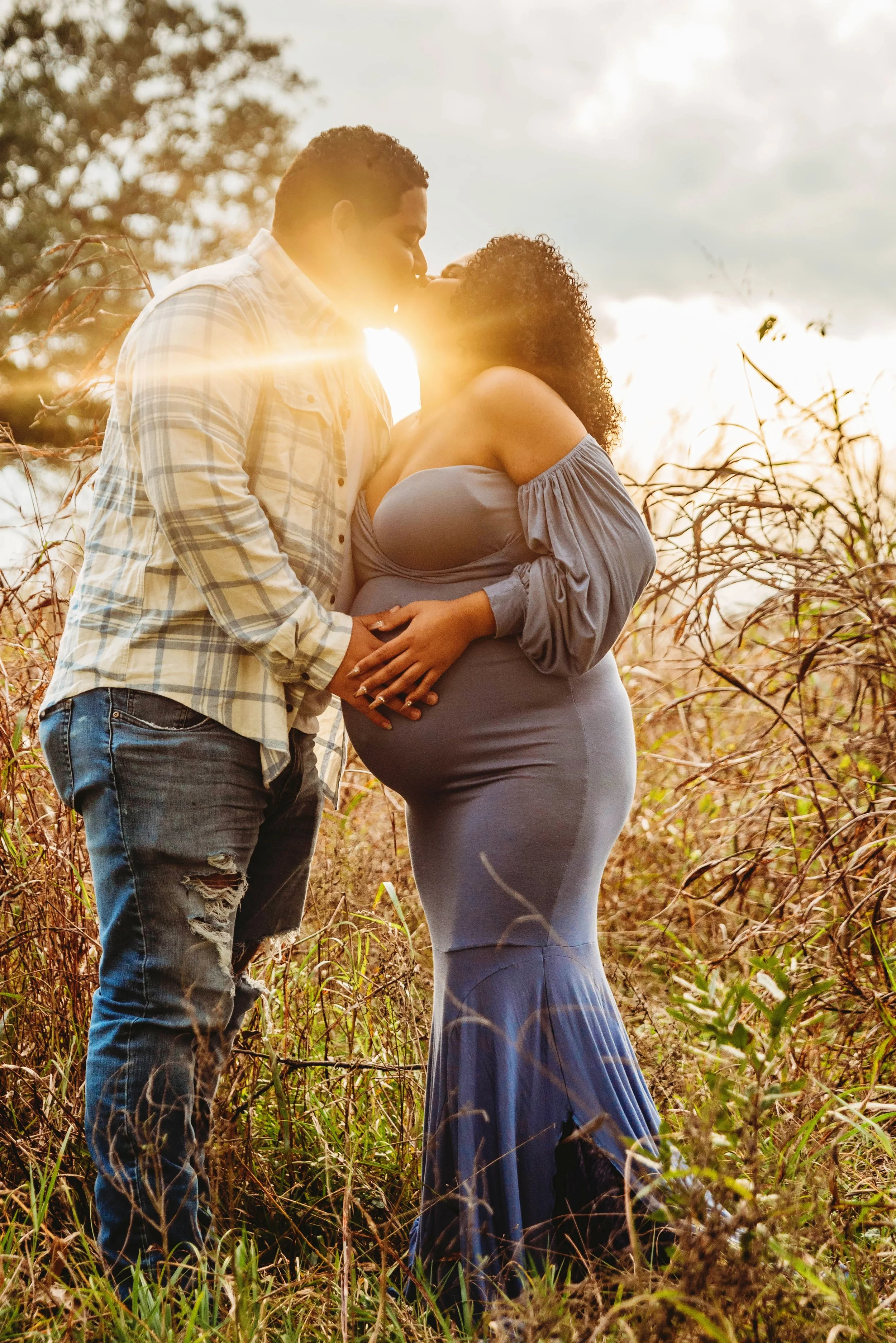 A couple sharing a romantic kiss outdoors during sunset, with the man holding the pregnant woman's belly in a field of tall grass.