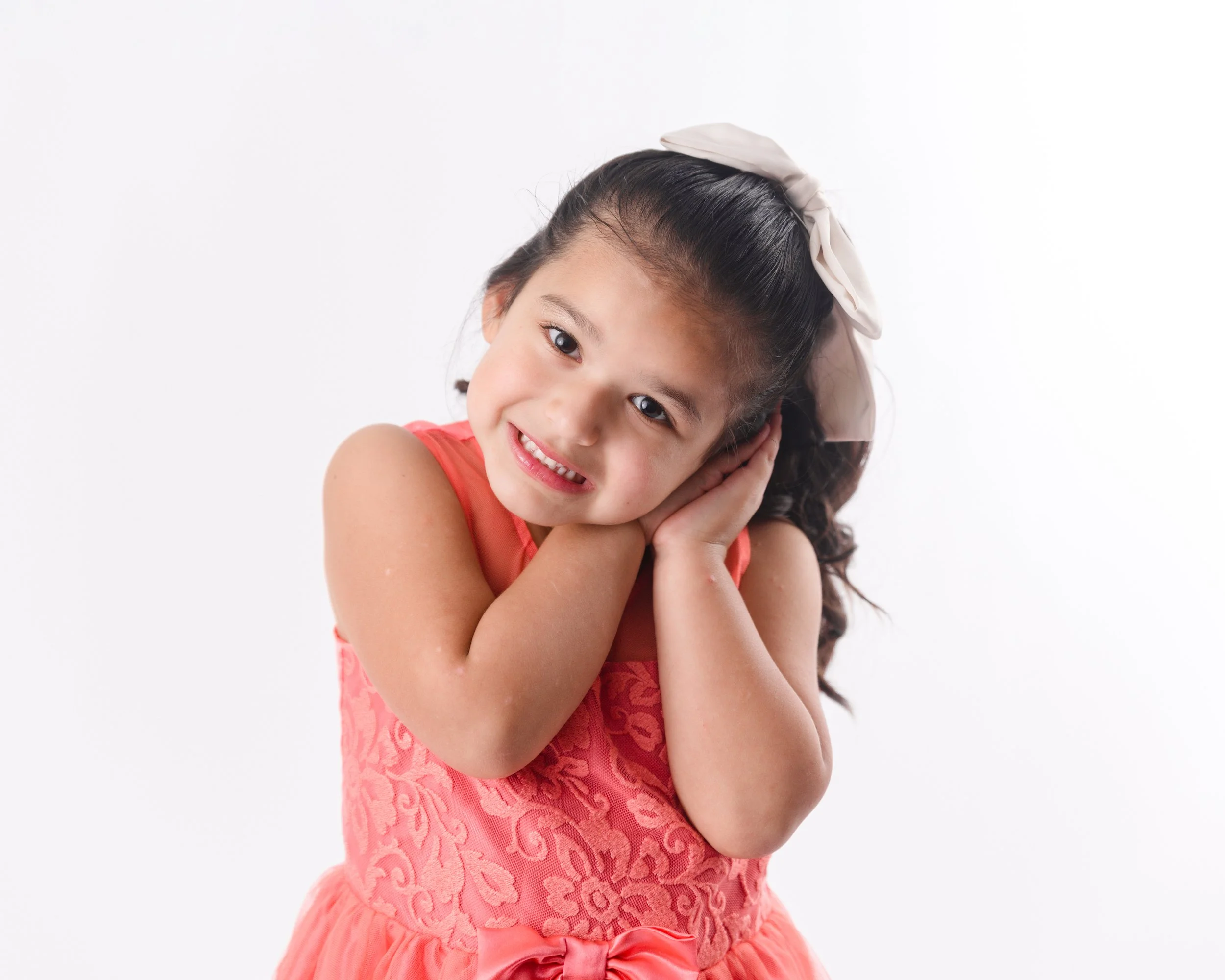 A young girl with dark hair, tied with a large white bow, wearing a coral dress with lace details and a bow at the waist, showing a sweet smile and gently touching her face with her hands.