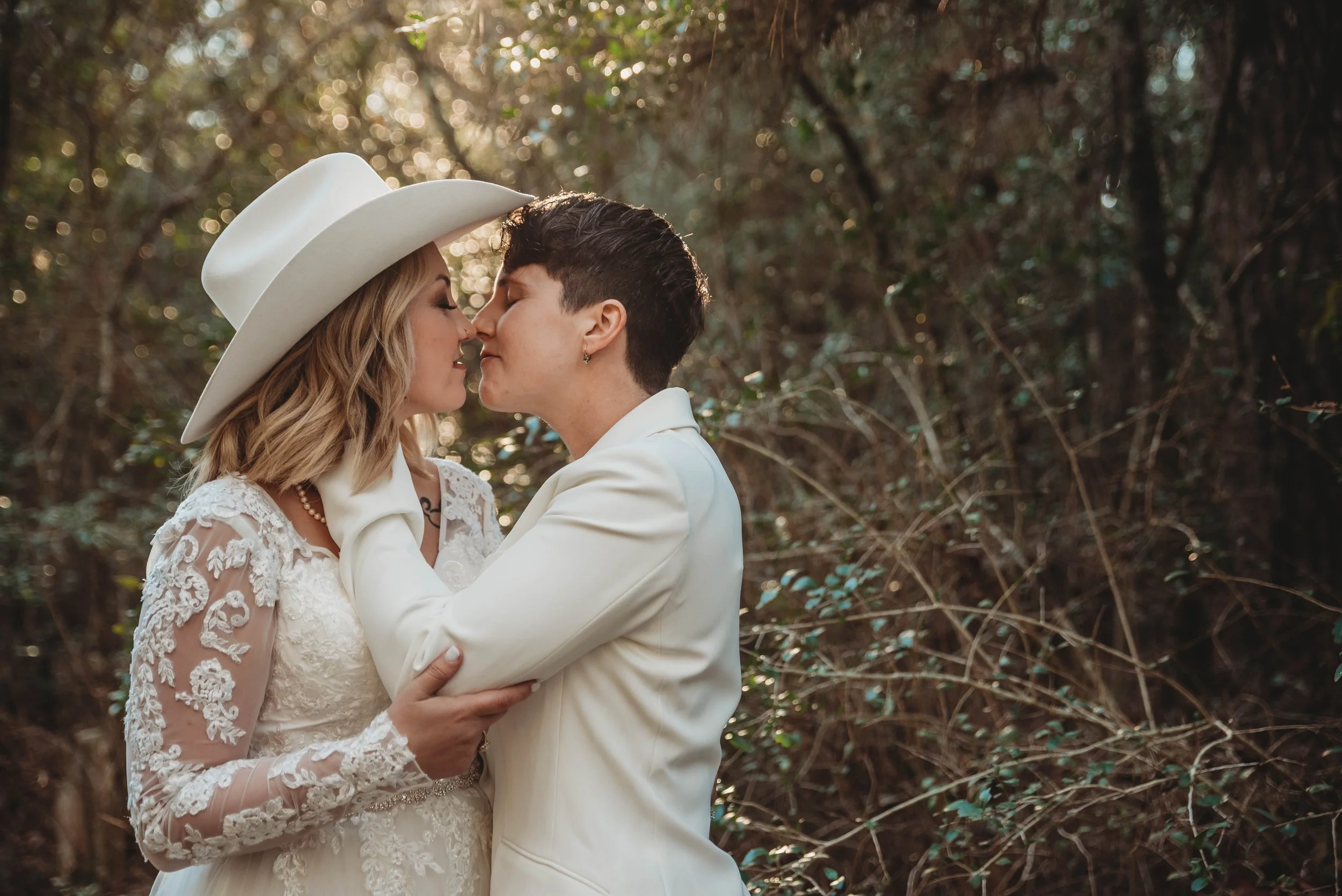 Two women, a bride in a lace wedding dress and a woman in a white suit, share a kiss outdoors surrounded by trees and sunlight.