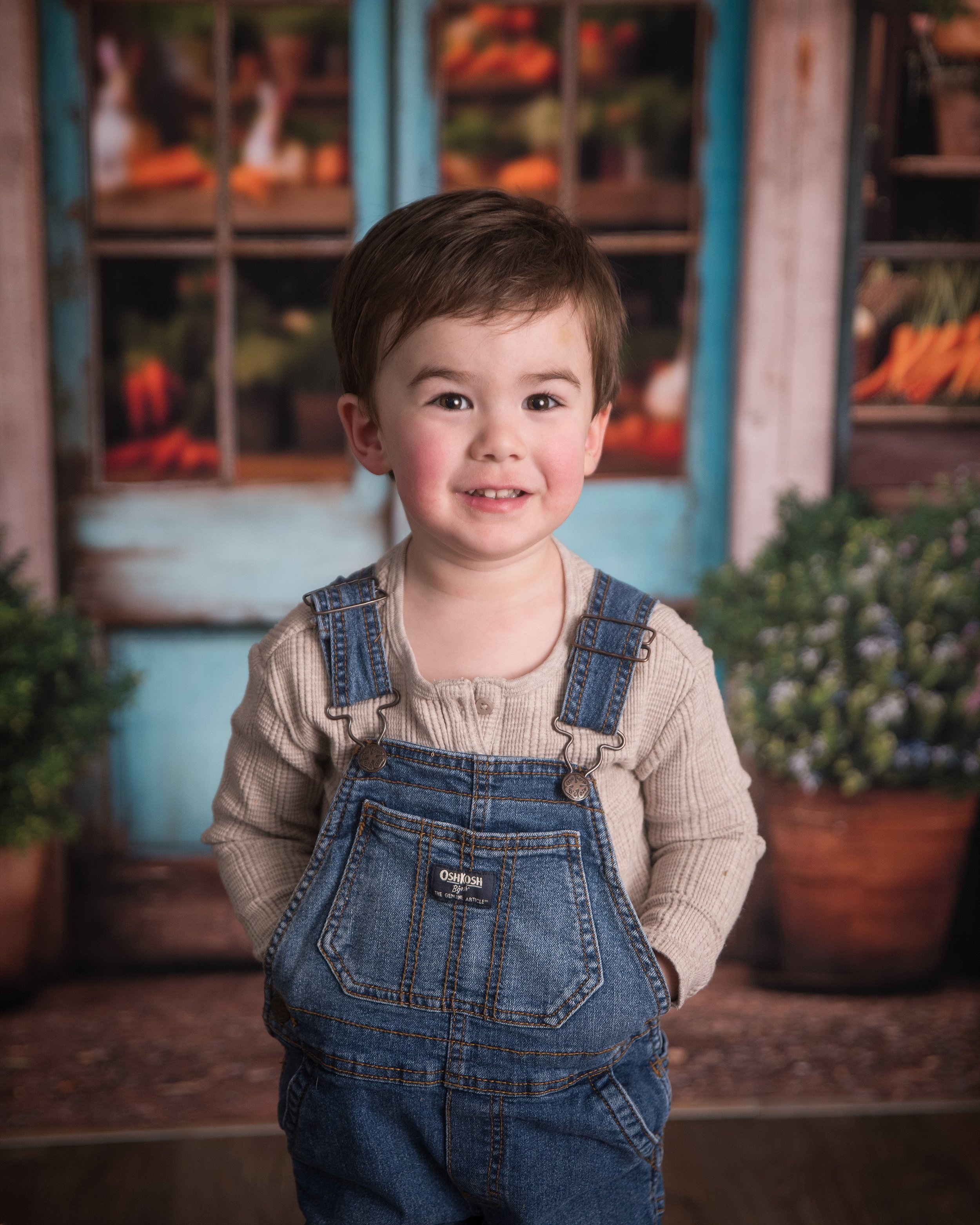 A young boy with brown hair, wearing a beige shirt and denim overalls, smiling outdoors with potted plants and a rustic background.