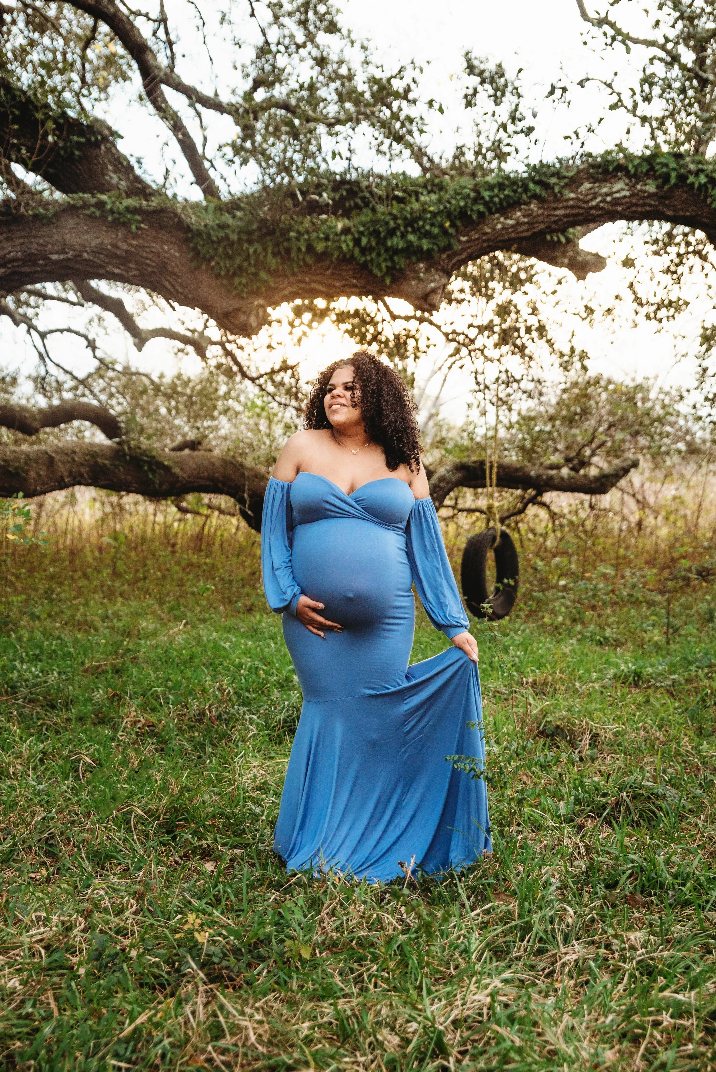 A pregnant woman in a blue dress stands outdoors next to a tree, smiling and gently holding her belly, with a tire swing hanging from the tree branches in the background.