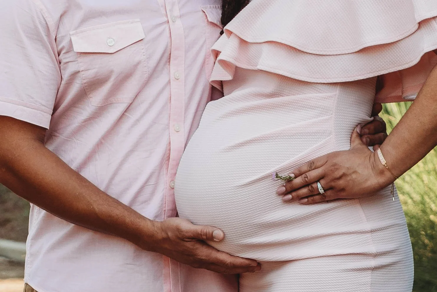 A couple with darker skin tones, the man wearing a light pink shirt and the woman wearing a light pink dress, embrace outdoors. The woman's pregnant belly is visible, and her hand rests on her belly.