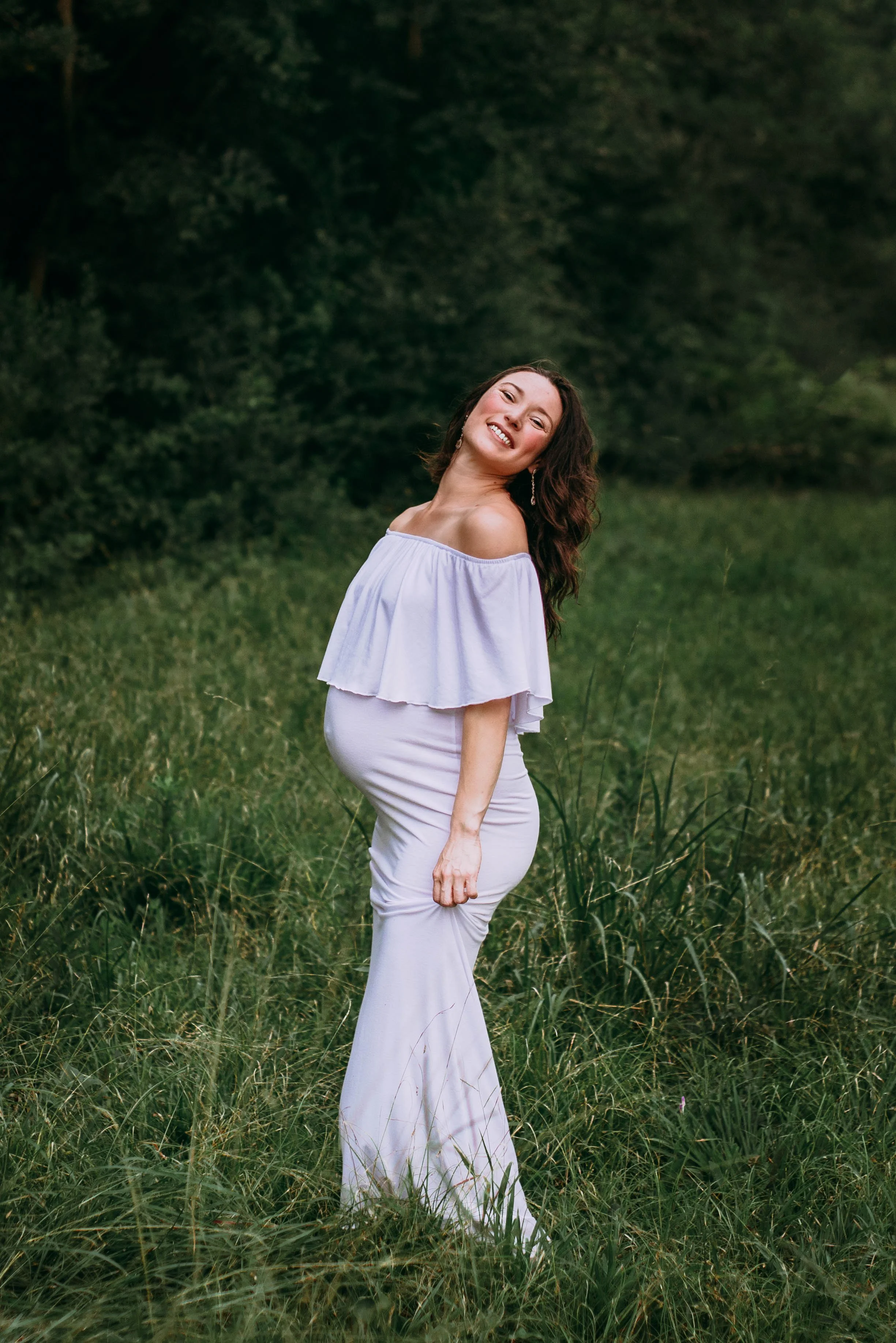 A pregnant woman with long, wavy brown hair smiling and posing outdoors in a grassy field with trees in the background, wearing a white off-the-shoulder dress.