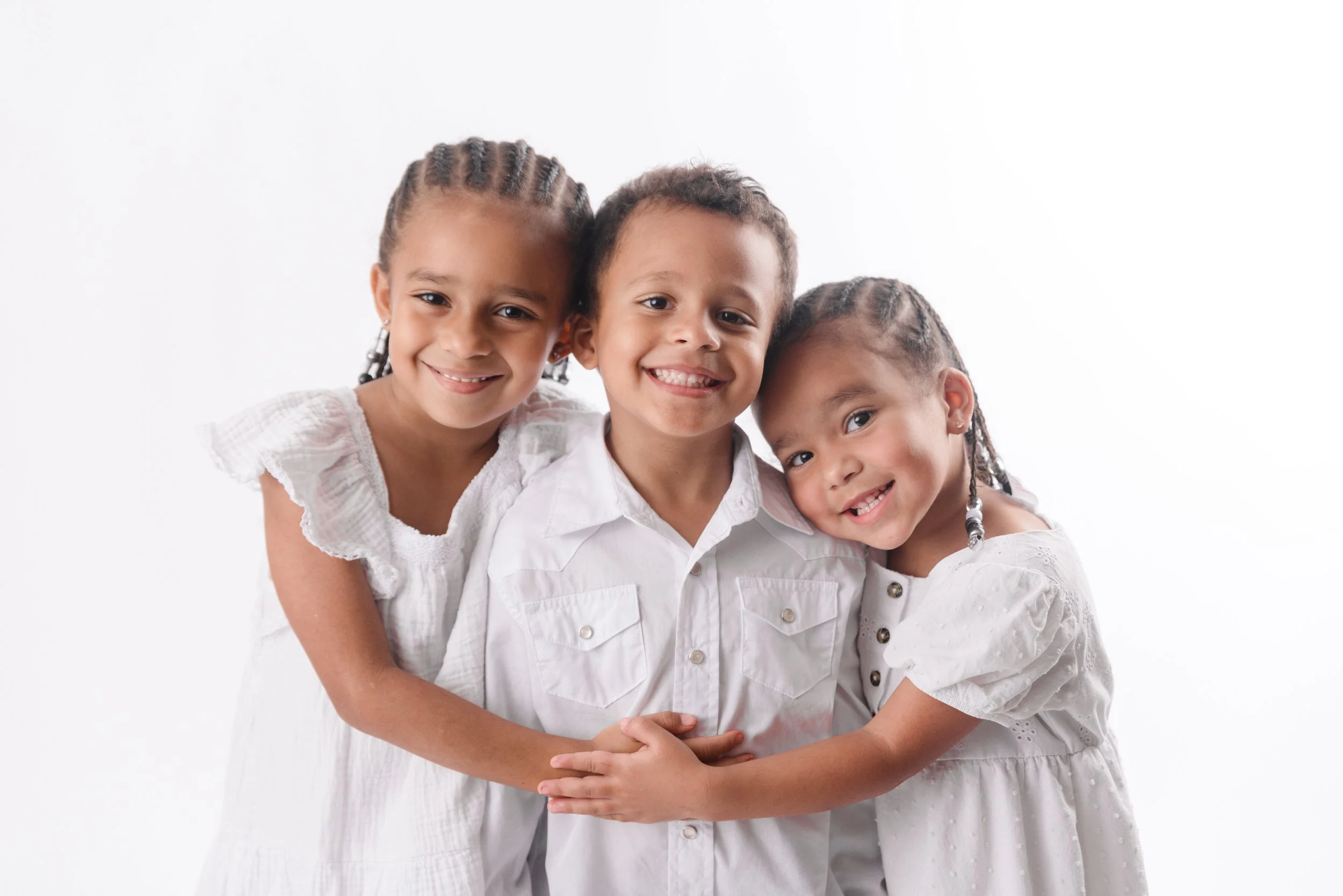 Three happy children hugging each other, wearing white clothing, with a white background.