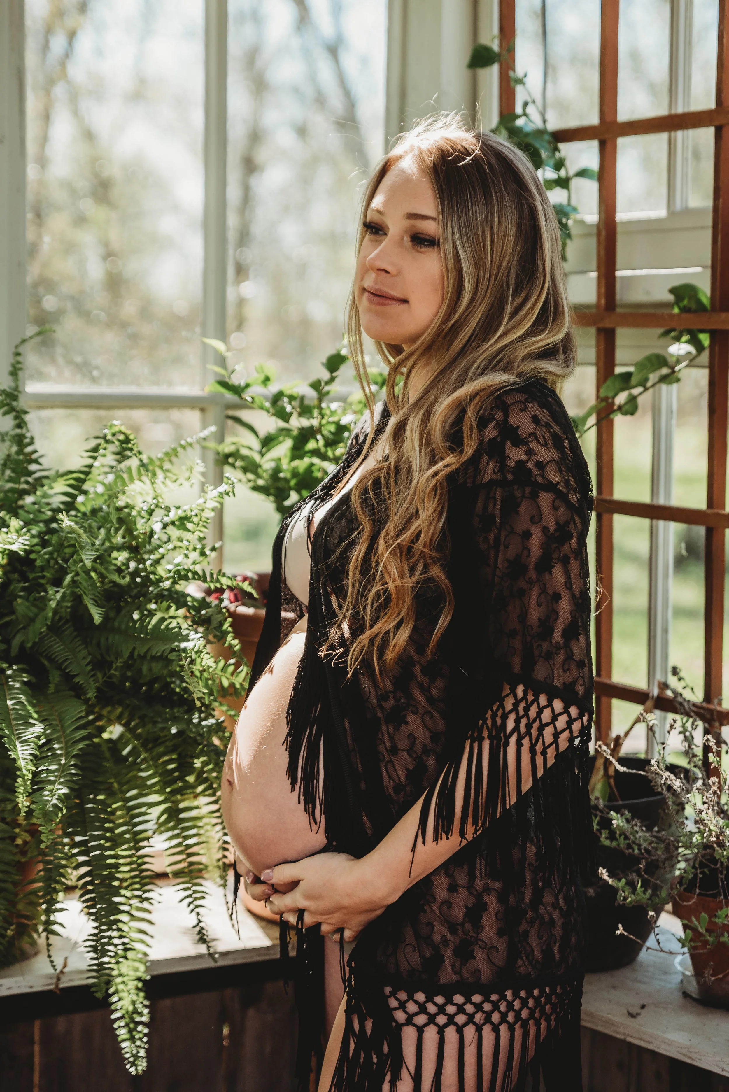 Pregnant woman with long wavy hair standing in a sunlit room with green plants, wearing a black lace cover-up.