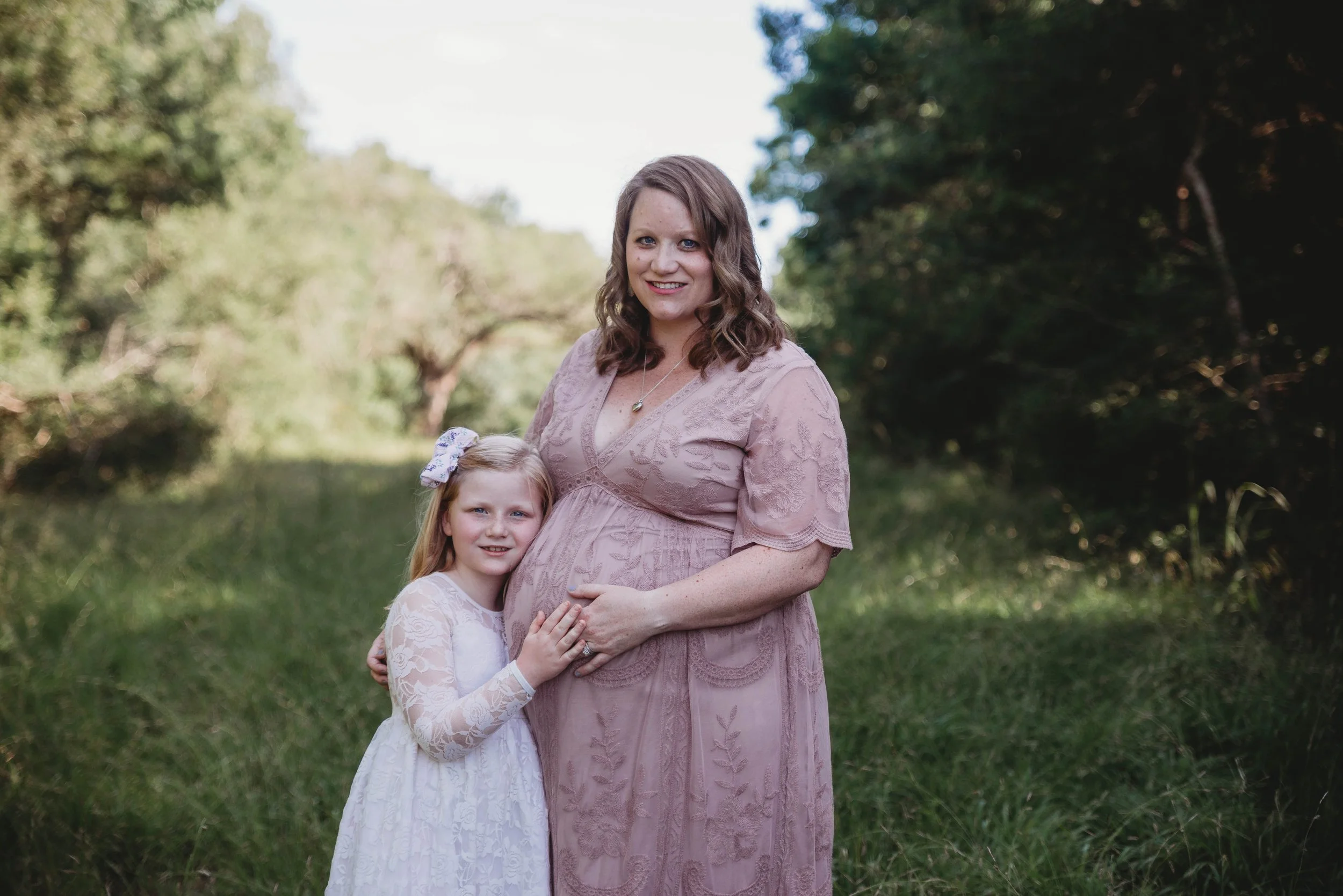 A pregnant woman in a pink lace dress standing outdoors, embracing a young girl in a white lace dress with a bow in her hair, on a grassy area with trees in the background.