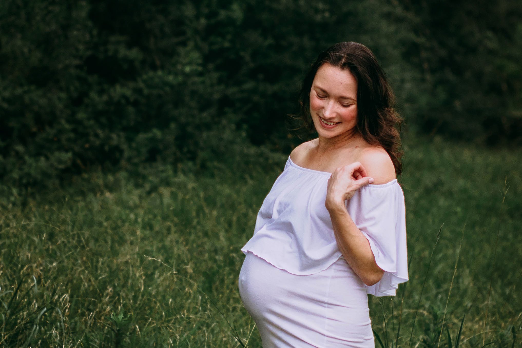 Pregnant woman standing in a grassy field, smiling and touching her shoulder, wearing a white off-the-shoulder dress.