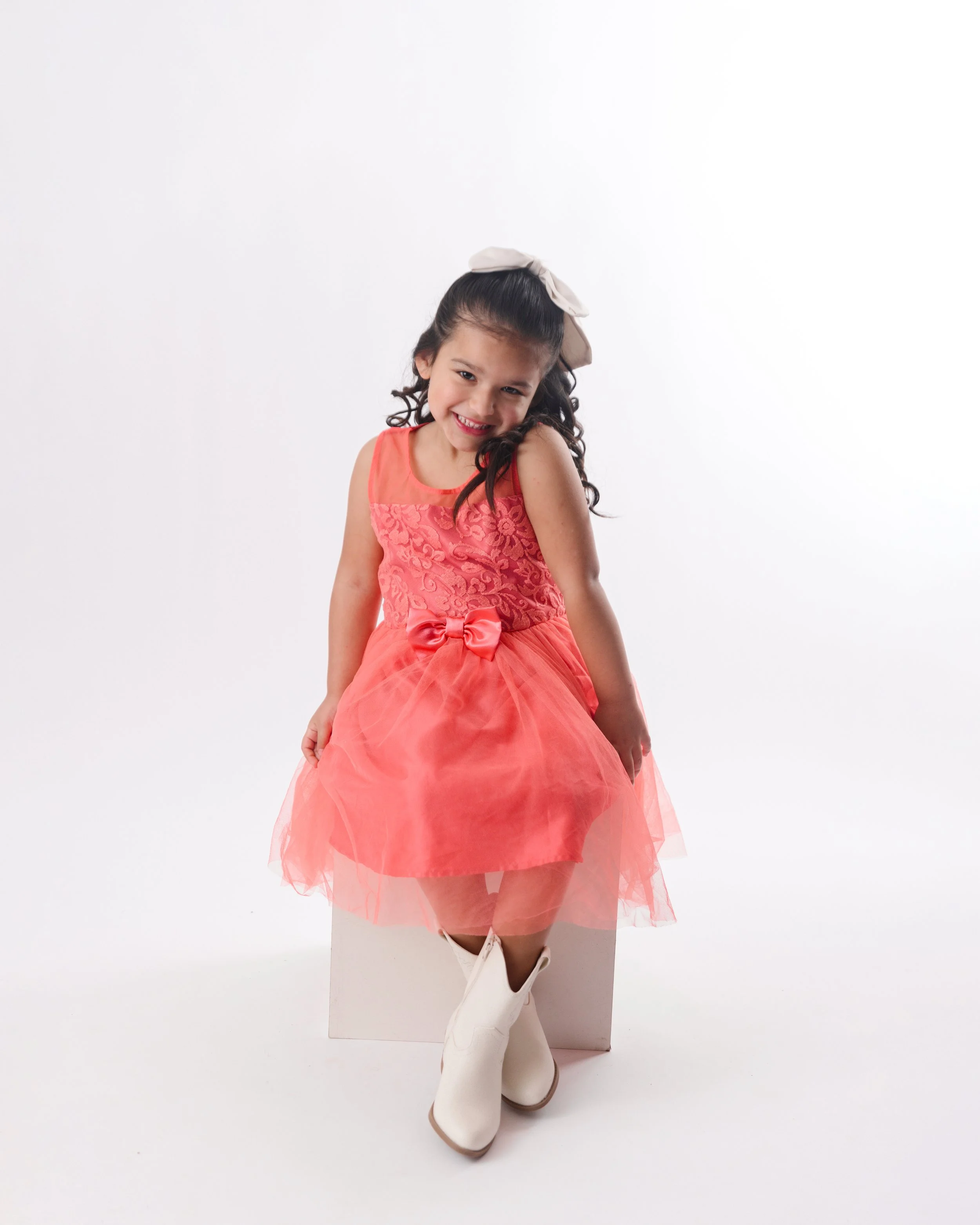 Young girl in a coral pink dress with a bow, white cowboy boots, sitting on a white cube against a white background, smiling.