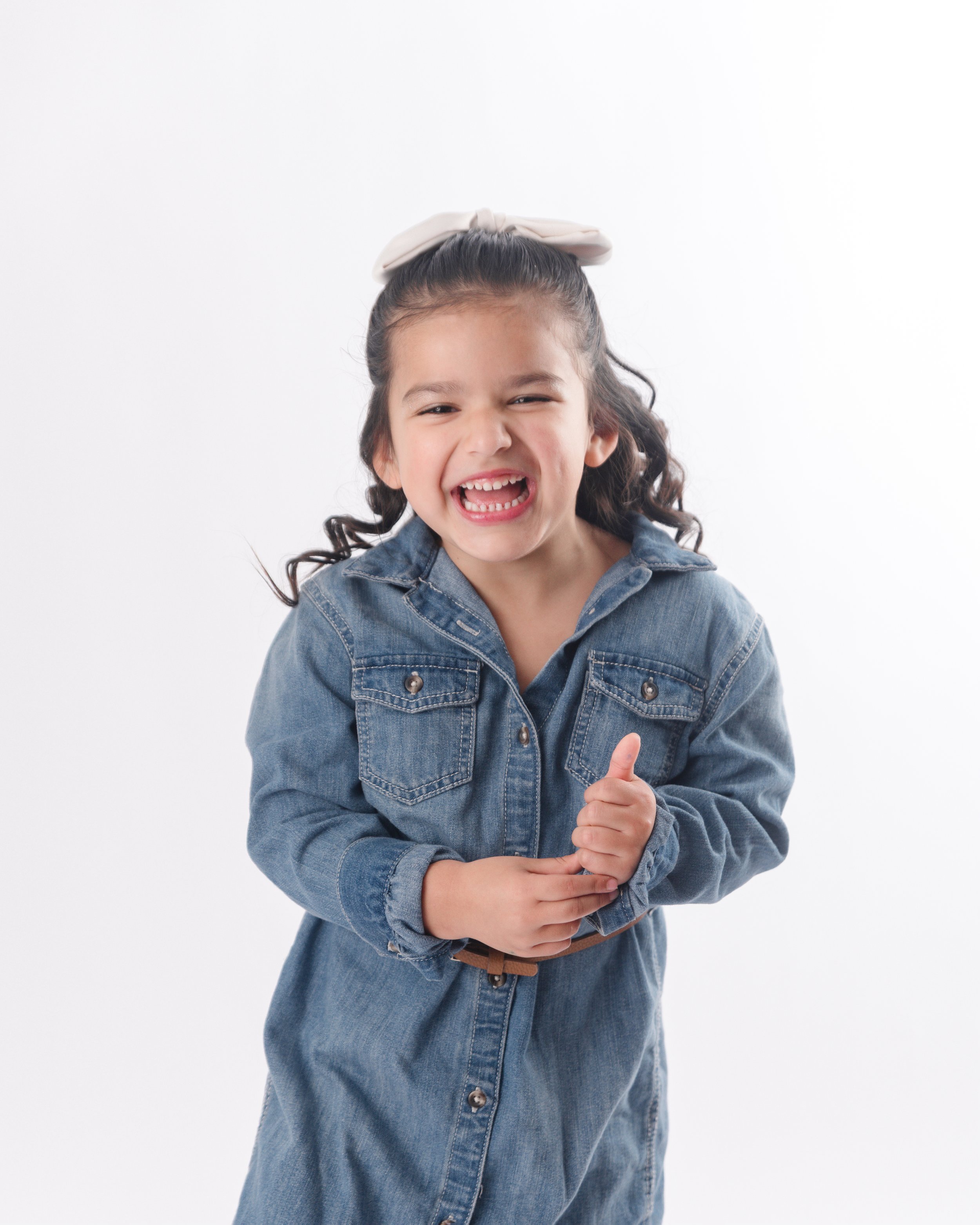 Smiling young girl with curly hair, wearing denim jacket and bow in hair, showing thumbs up.