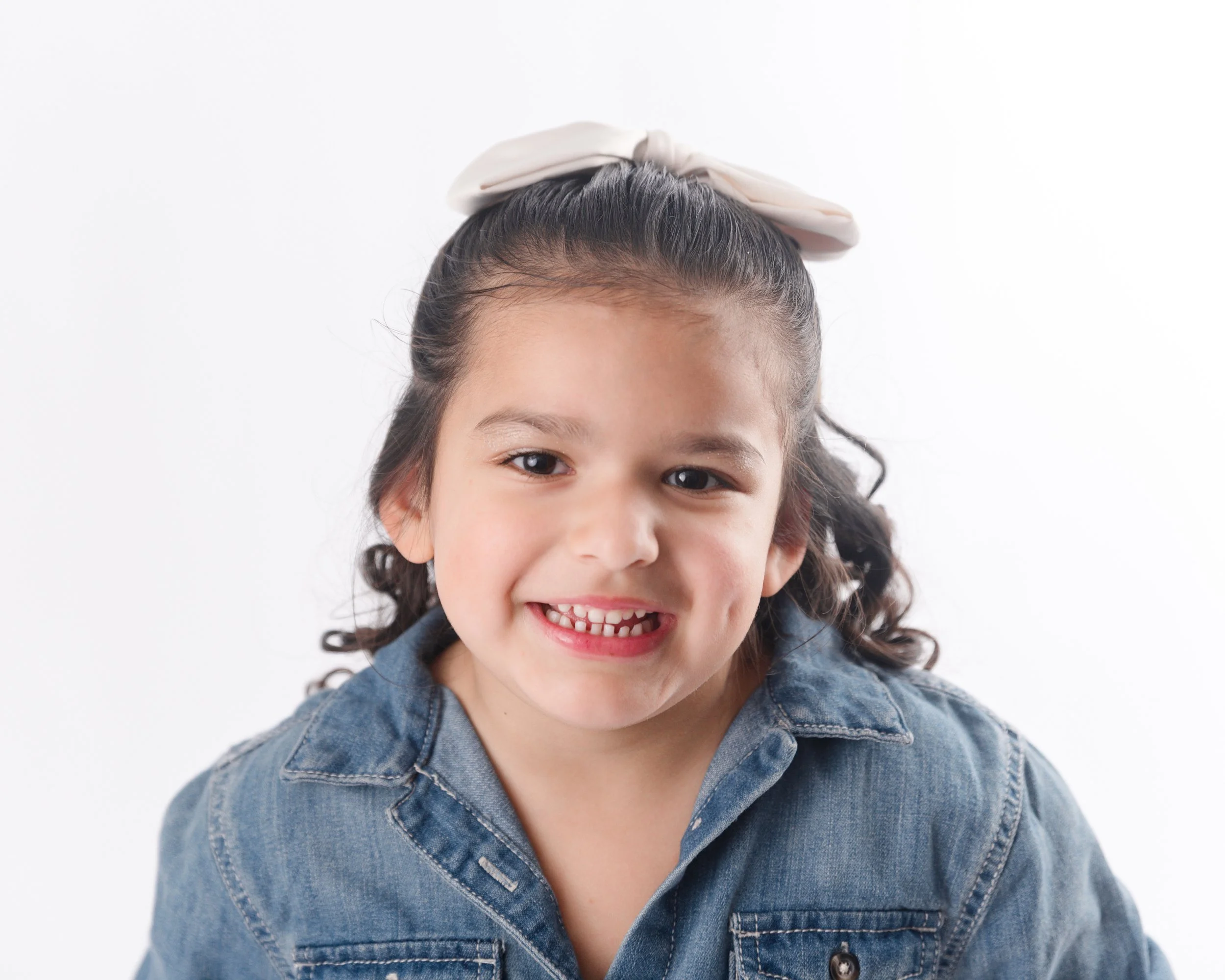 A young girl with long, curly hair tied with a white bow, wearing a denim jacket, smiling at the camera.