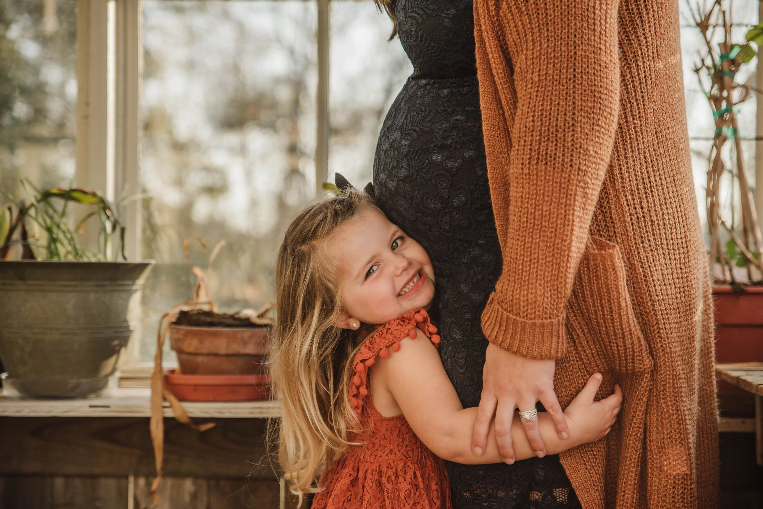 A young girl hugging a pregnant woman, smiling at the camera, indoors with houseplants and a window in the background.