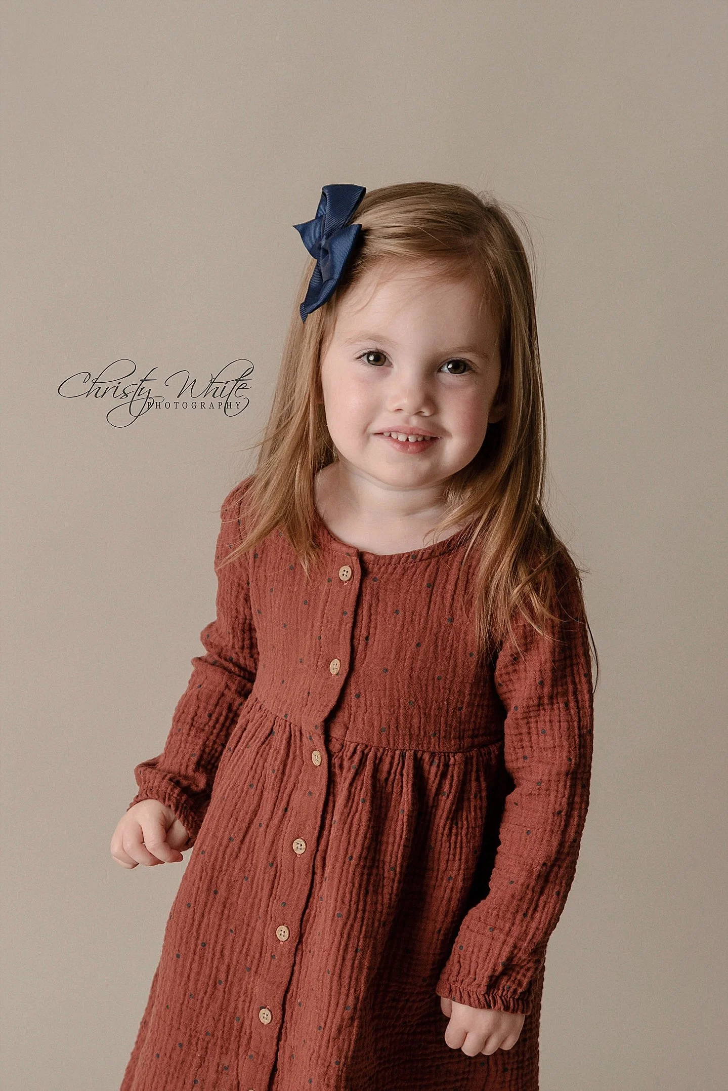 A young girl with red hair wearing a rust-colored dress with small black dots and a navy blue hair bow, smiling softly at the camera.