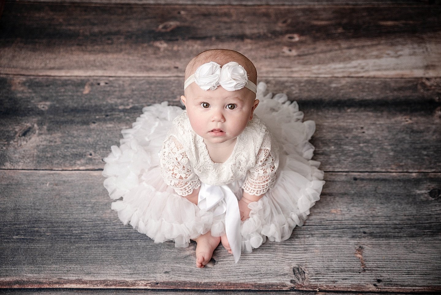 A baby girl dressed in a white lace dress and tutu, with a matching headband, sitting on a wooden floor and looking up at the camera.