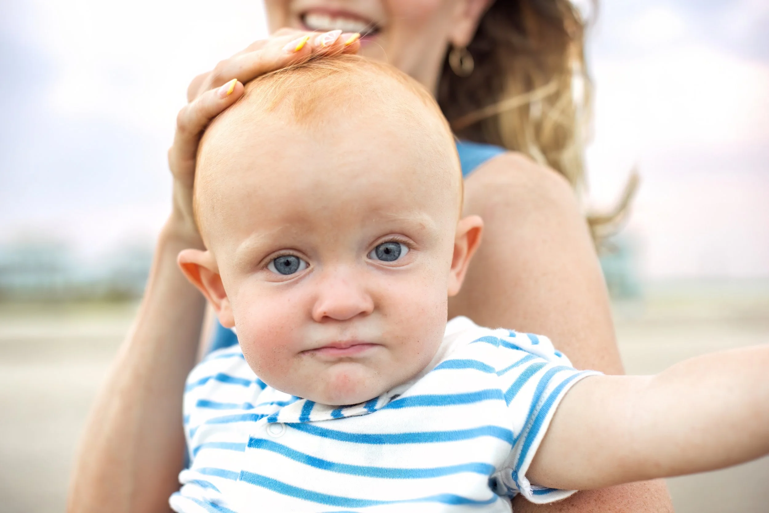 Close-up of a young child with blue eyes and a serious expression, being held by an adult with blonde hair, outdoors on a bright day.