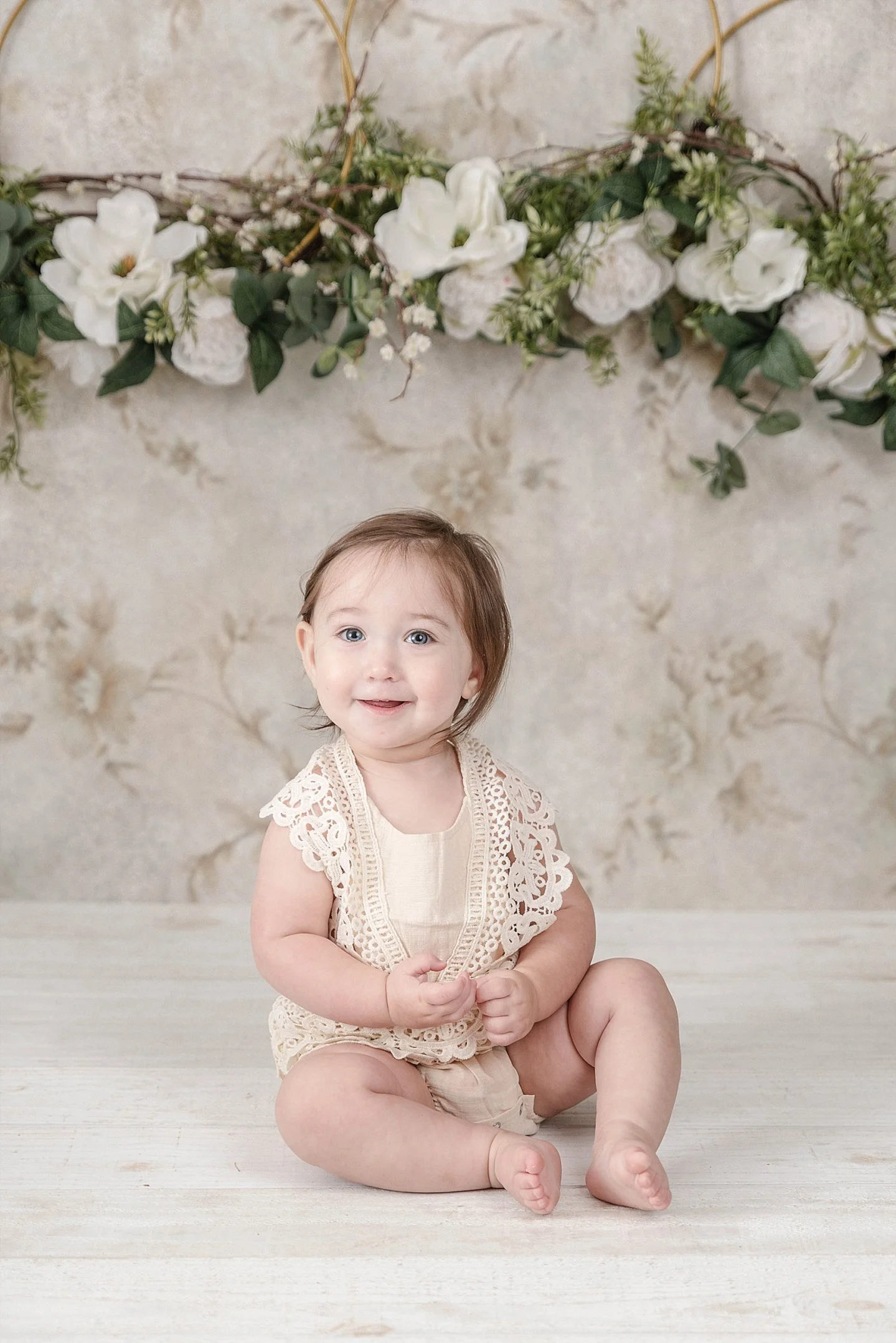 A smiling young toddler girl sitting on a light-colored wooden floor with a floral garland hanging against a textured wall behind her.