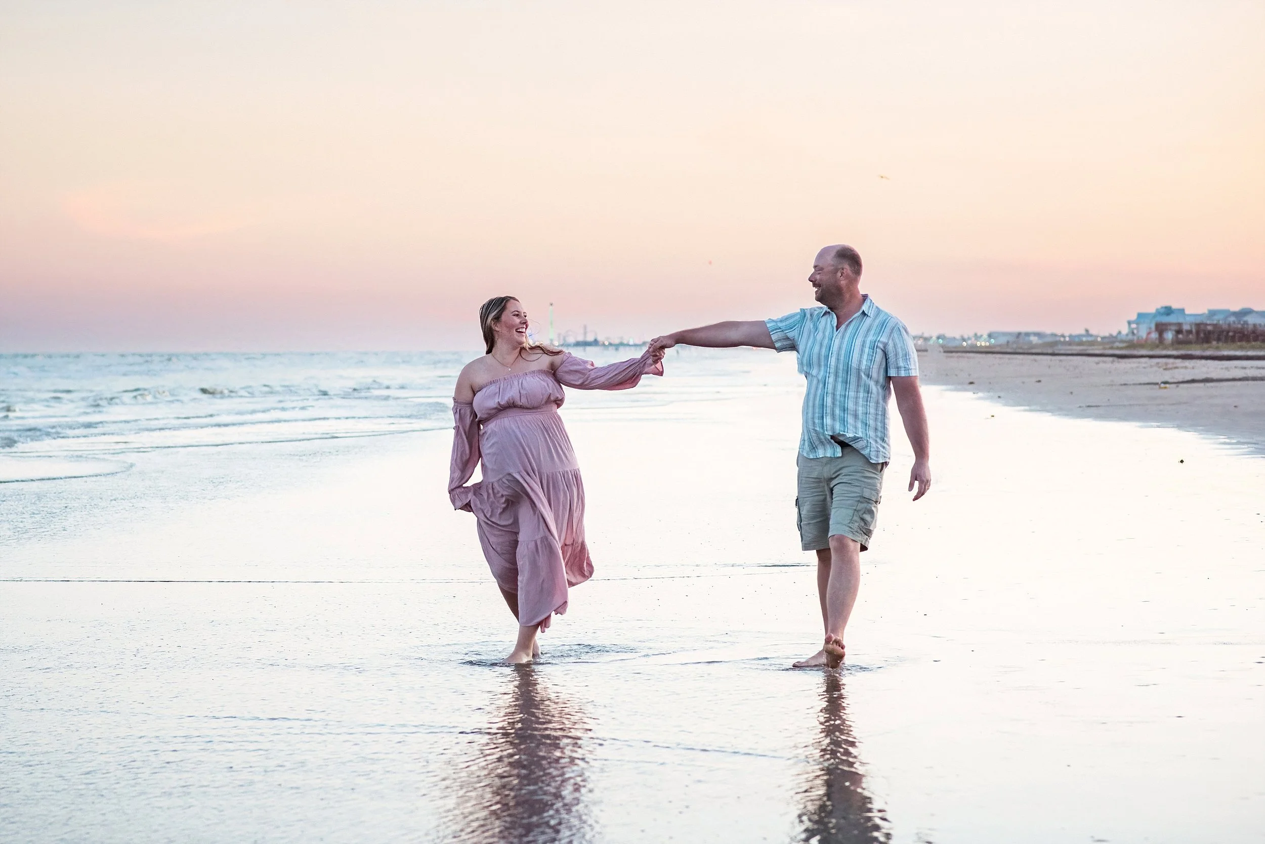 Expecting mother wearing a soft pink maternity gown walking along the shoreline during golden hour in Galveston for a romantic beach maternity session.