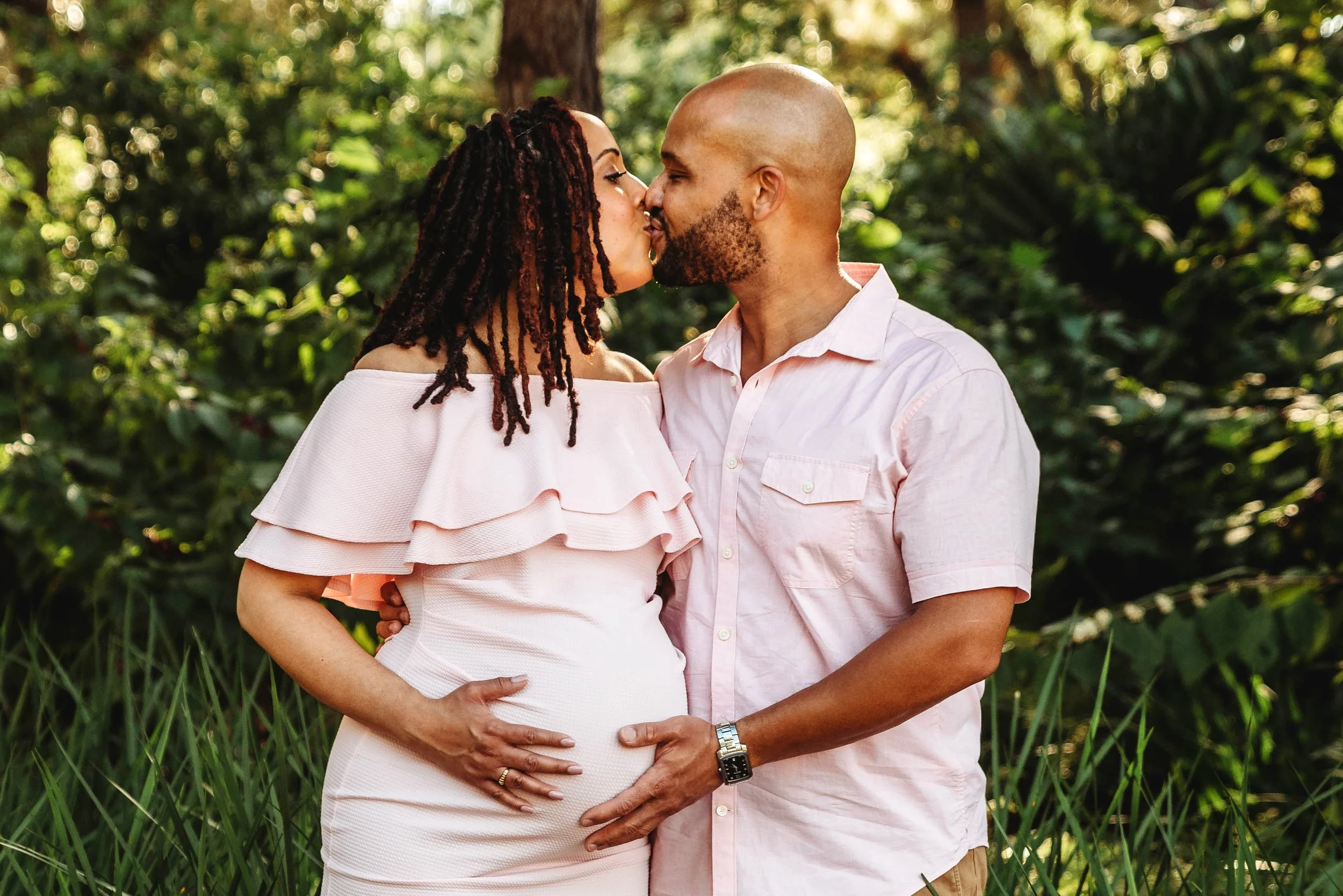 A pregnant woman and a man sharing a kiss outdoors, with lush green foliage in the background.