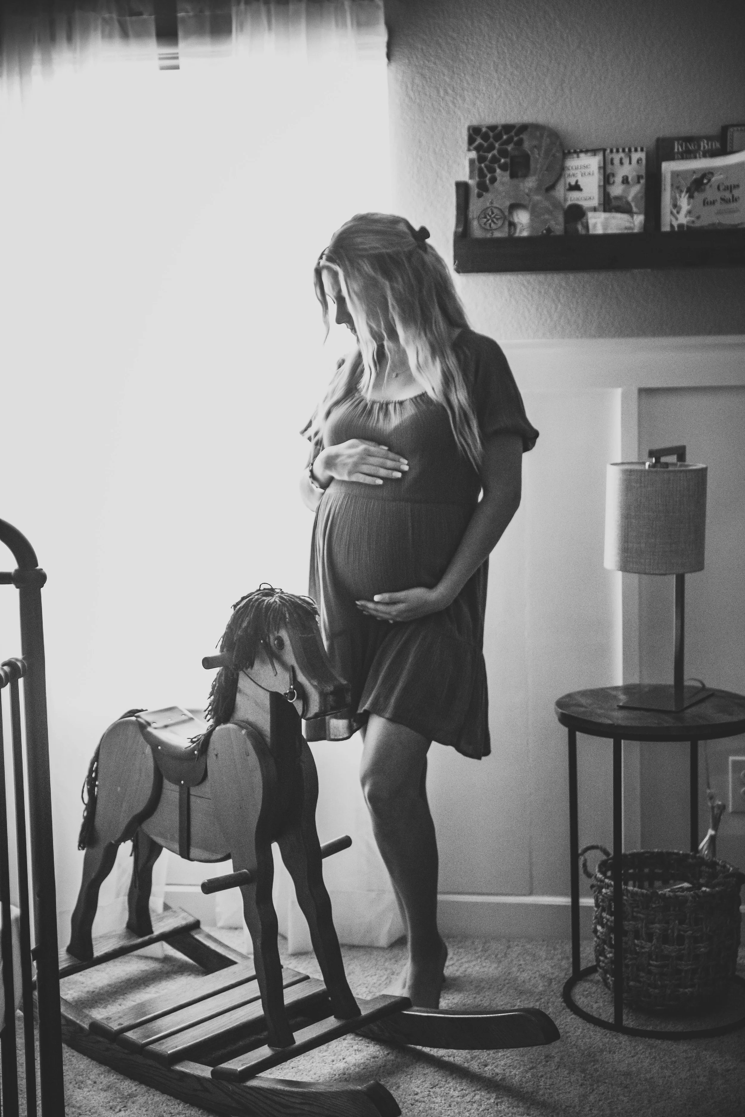 A pregnant woman standing indoors next to a rocking horse, holding her belly with one hand and resting another hand on her stomach, near a window with bright light. The room has a side table with a lamp, a basket, and shelves with books.