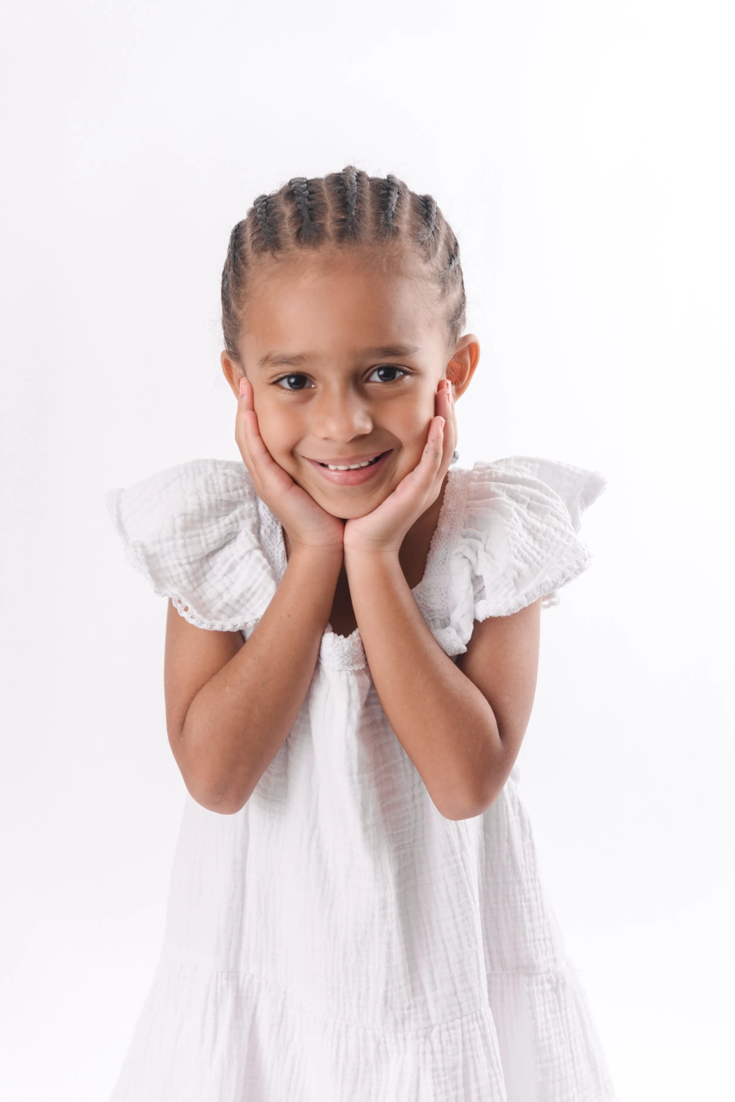 A young girl with braided hair wearing a white dress, smiling and resting her face in her hands against a plain white background.