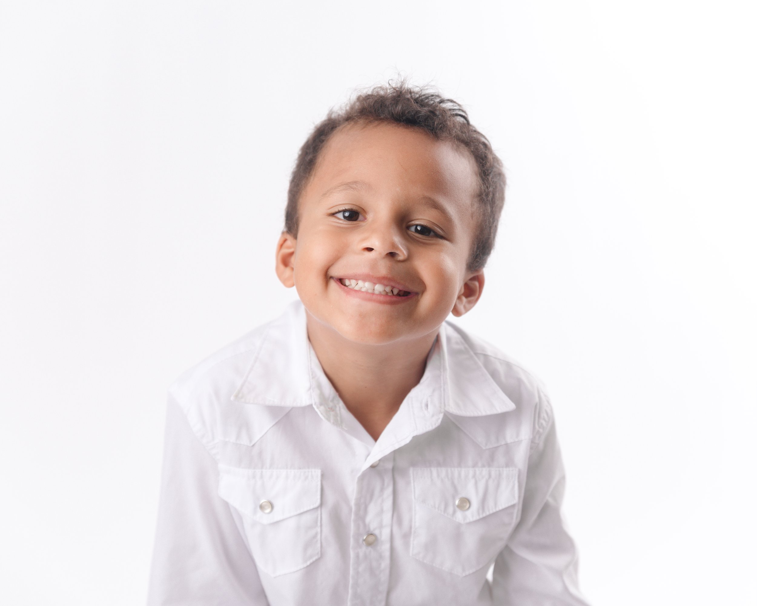 Smiling young boy with short dark curly hair, wearing a white button-down shirt, standing against a plain white background.