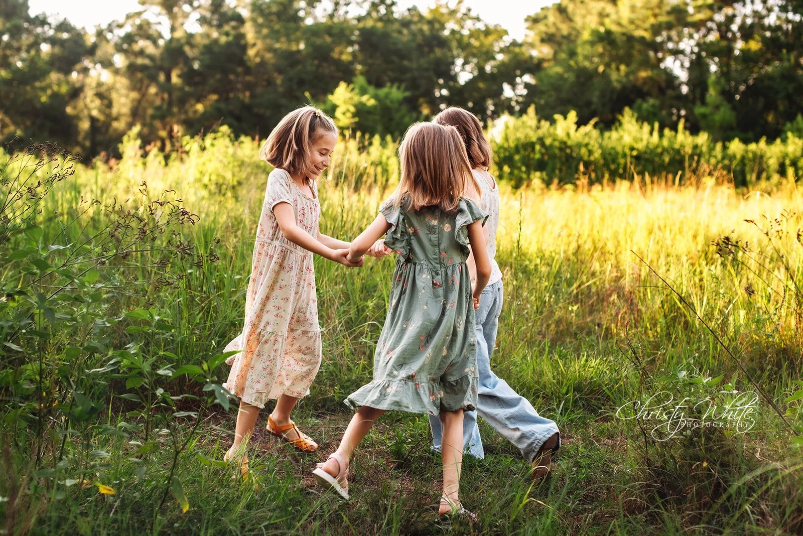 Four young girls holding hands and walking through a grassy field on a sunny day.