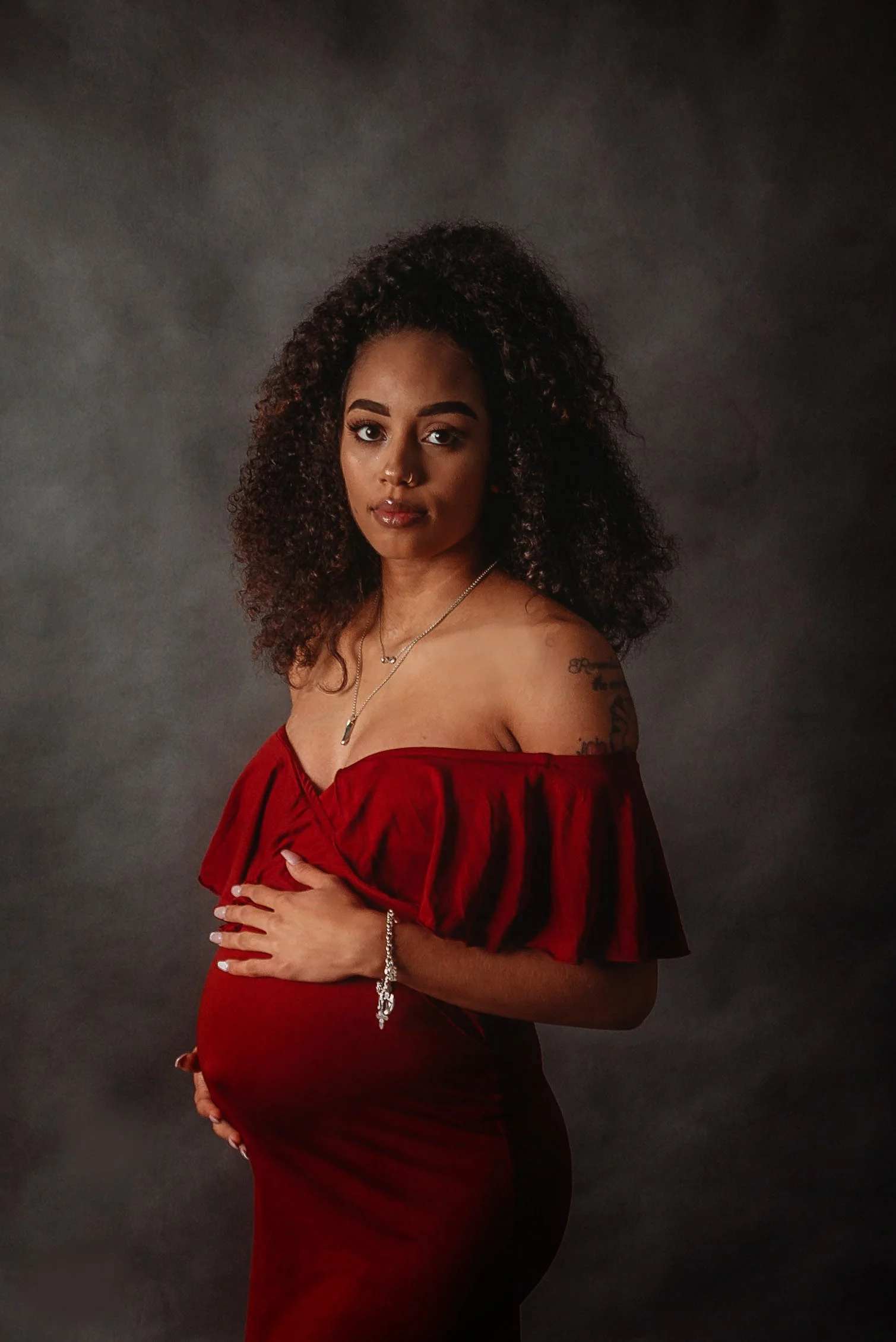 A pregnant woman with curly hair wearing a red off-the-shoulder dress, jewelry, and posing against a gray background.