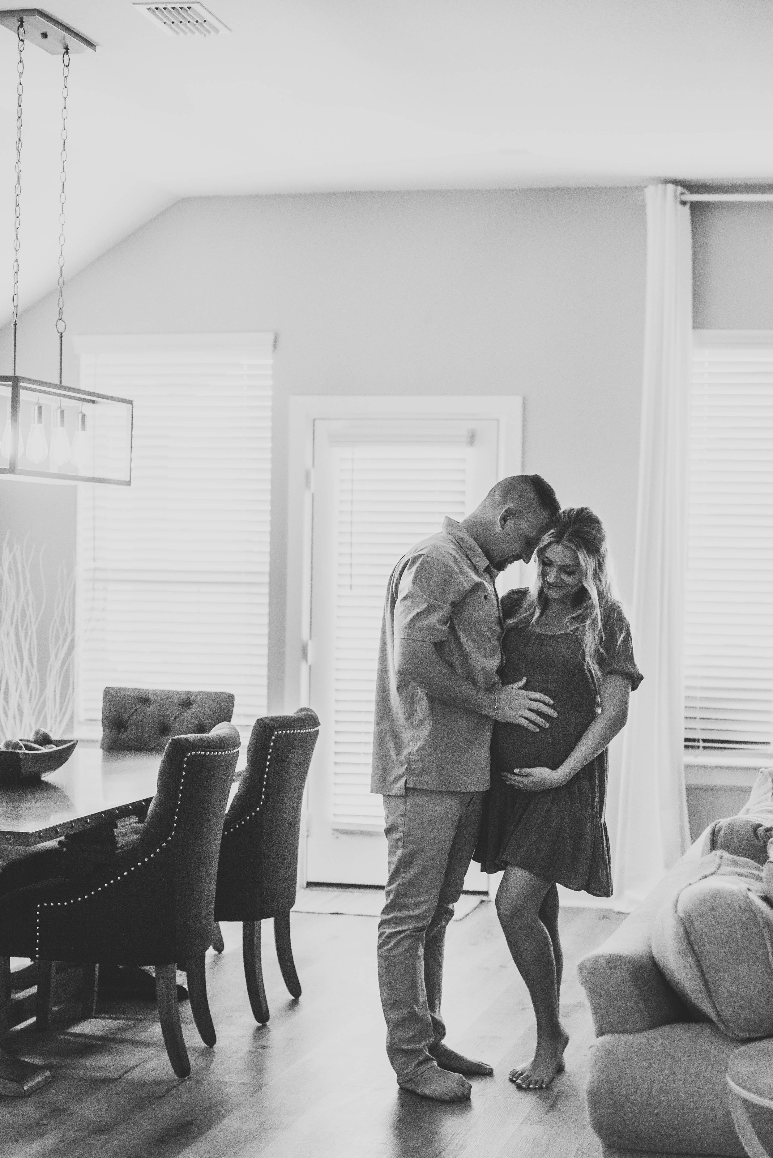 A couple standing close together in a living room, with the woman pregnant, and their foreheads touching gently.