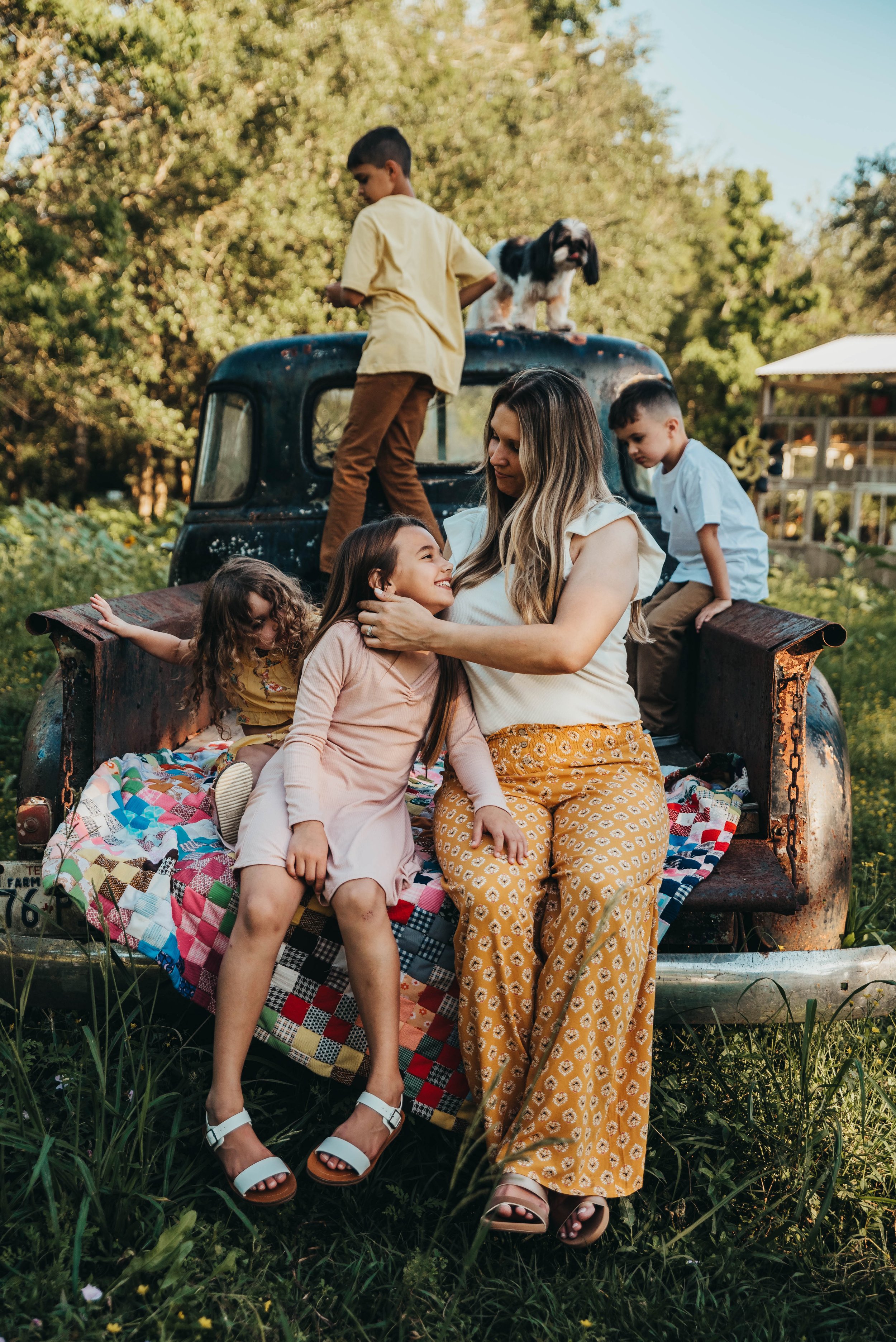 A woman and four children sitting and standing on the back of an old rusted truck, surrounded by green grass and trees, with a puppy and a boy standing on top of the truck.