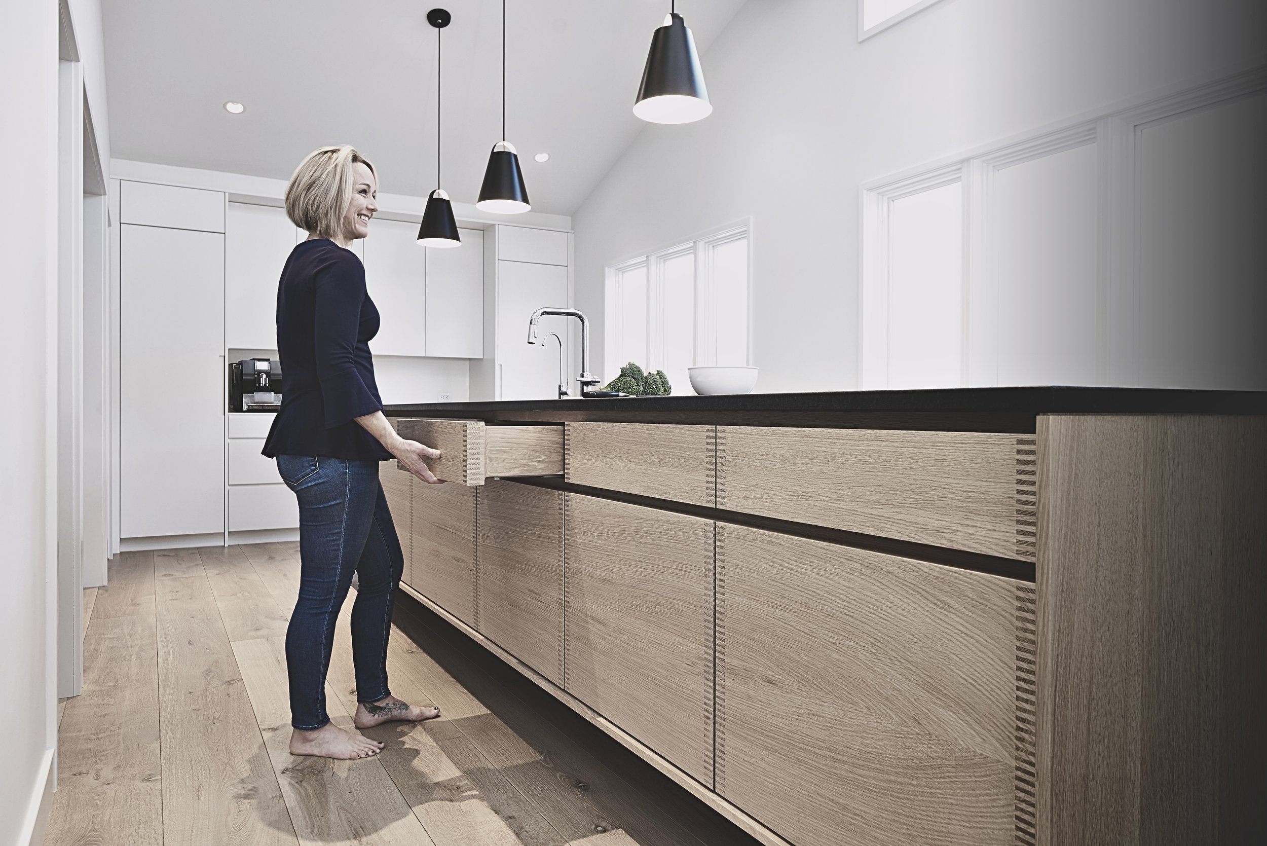 Woman in a blue blazer and jeans standing barefoot in a modern kitchen with light wood cabinets, black countertop, and pendant lights, pulling out a drawer from the island.