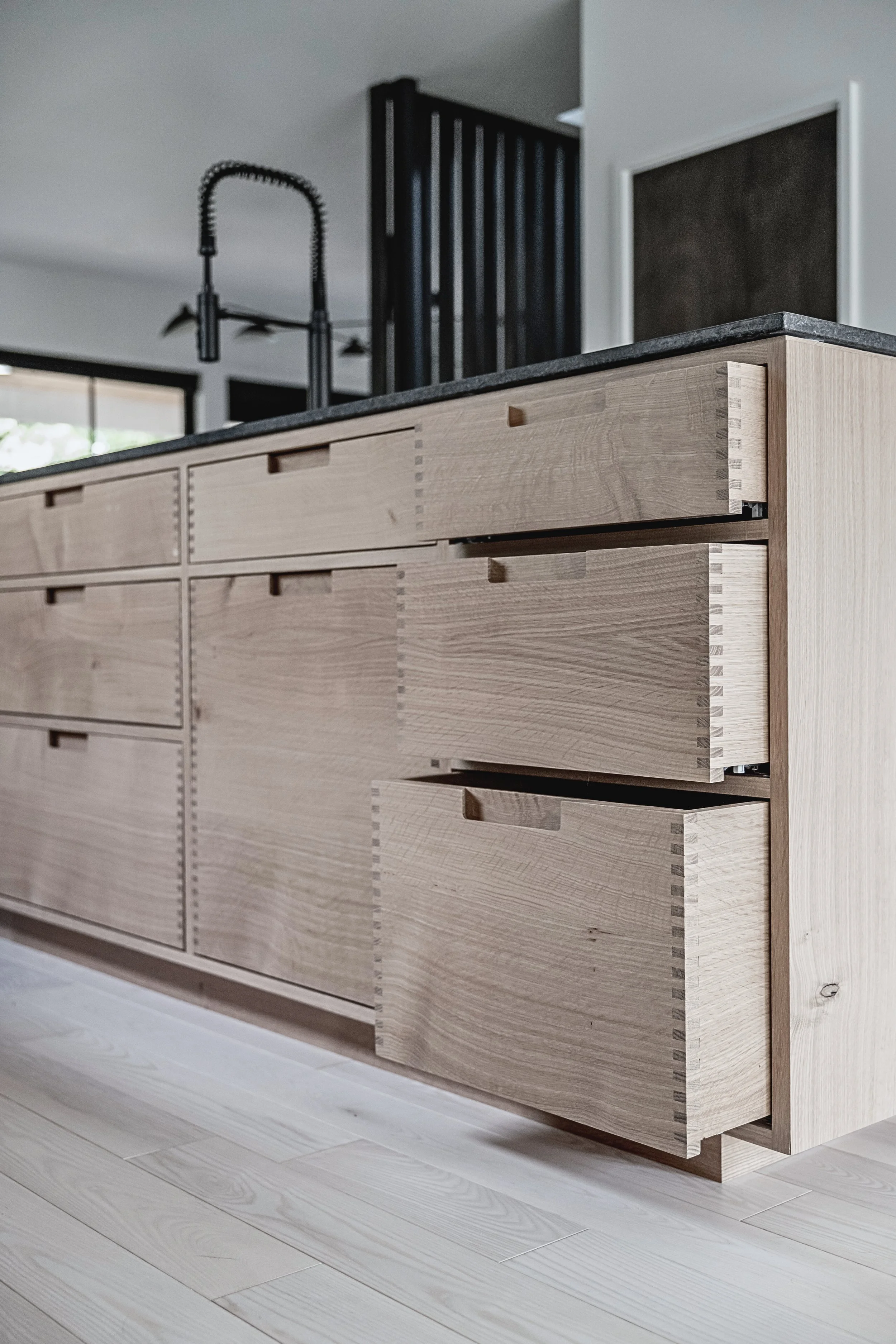 Close-up of a modern light wooden kitchen island with drawers in a bright kitchen, featuring a dark countertop and a black gooseneck faucet.