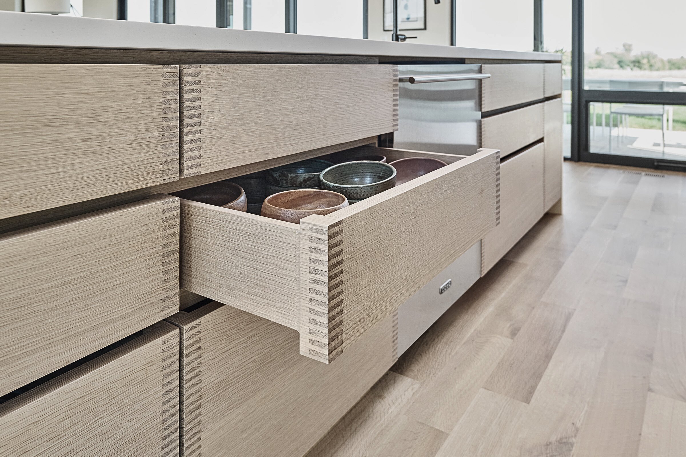 Open kitchen drawer with ceramic bowls inside, near a large window and wooden floor.