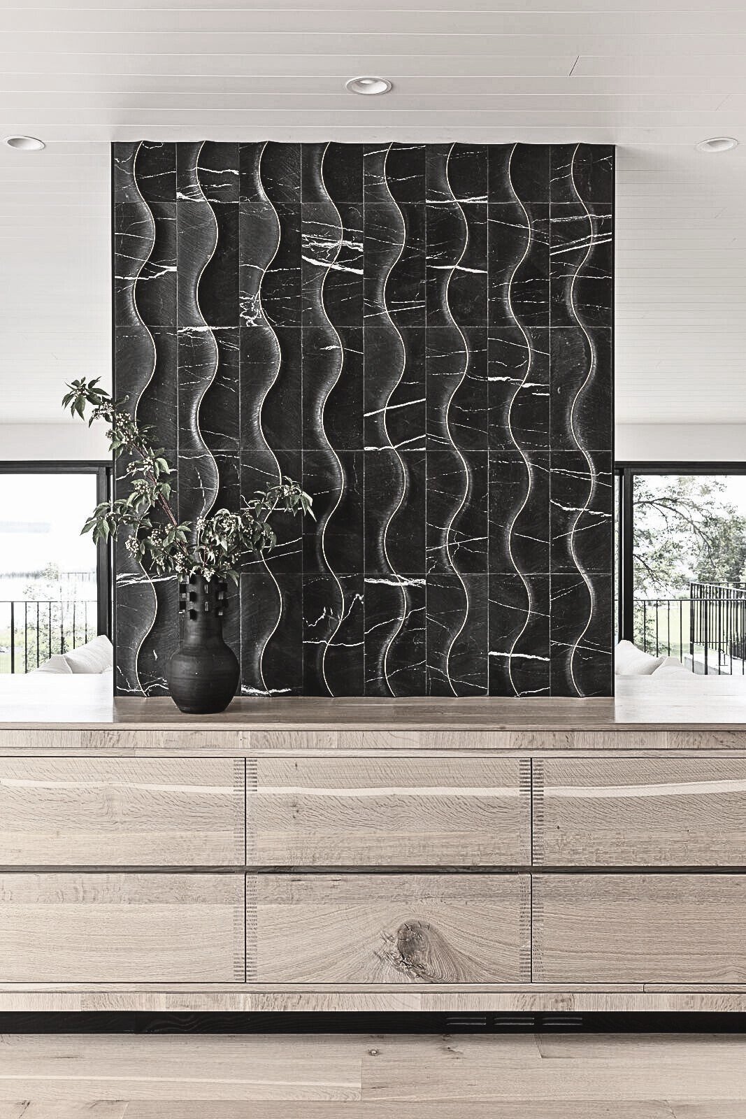 Modern interior with a large black marble accent wall with wavy white veins, a wooden sideboard, and a black vase with greenery on top. Two windows with balcony railings are visible, and a white ceiling with recessed lighting.