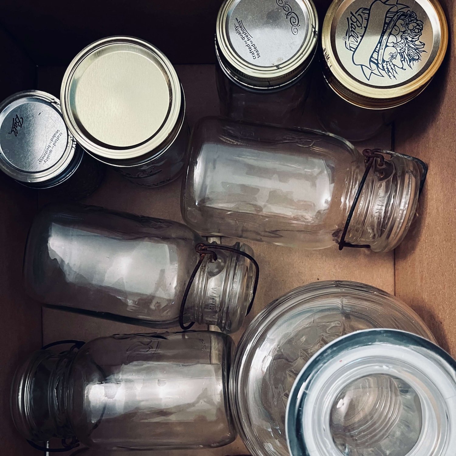 A top-down view of a cardboard box filled with vintage glass jars, including clear wire-bail mason jars with rusty metal clamps and several upright Ball canning jars with silver and gold lids, in a Greensboro, North Carolina home.