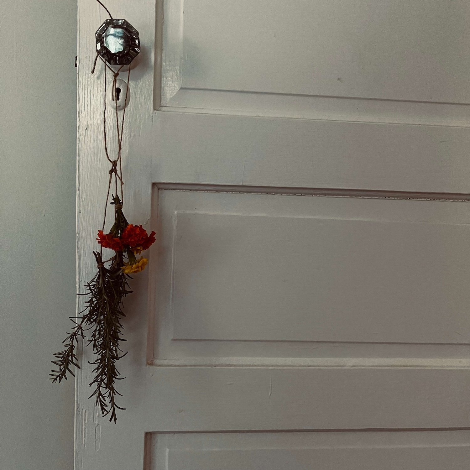 A hand-tied bundles of rosemary sprigs and orange and yellow marigold flowers hanging by a simple piece of twine from a vintage crystal doorknob on a white paneled interior door of a Greensboro bungalow.