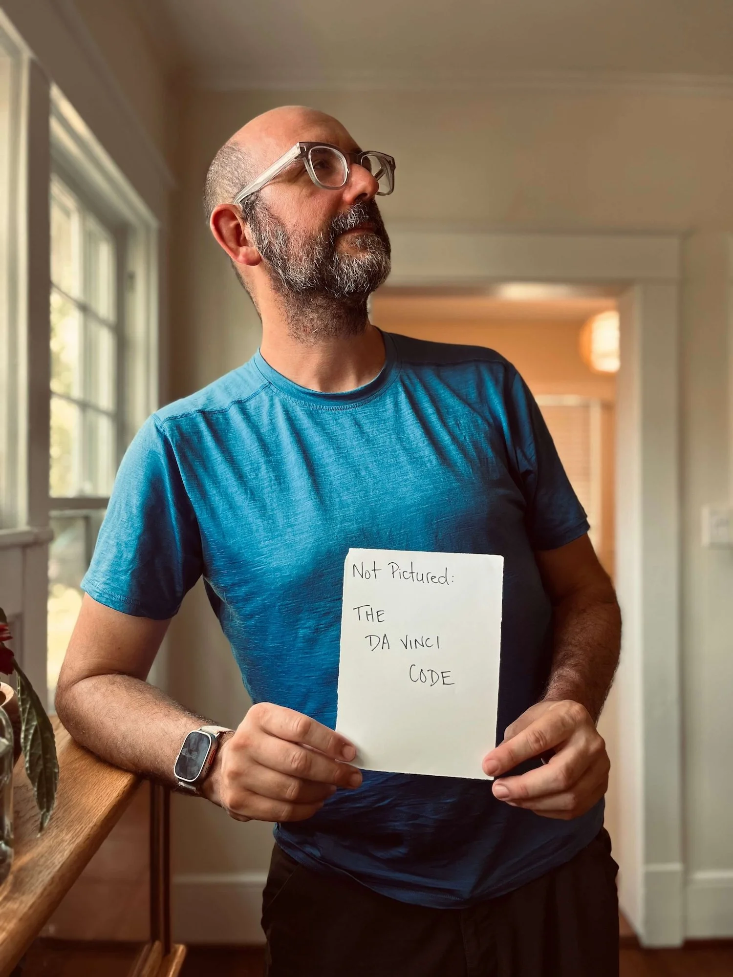 A candid, humorous portrait of a man with glasses and a beard leaning against a wooden counter inside a bright craftsman bungalow, holding up a small hand-written note that reads "Not Pictured: The Da Vinci Code."