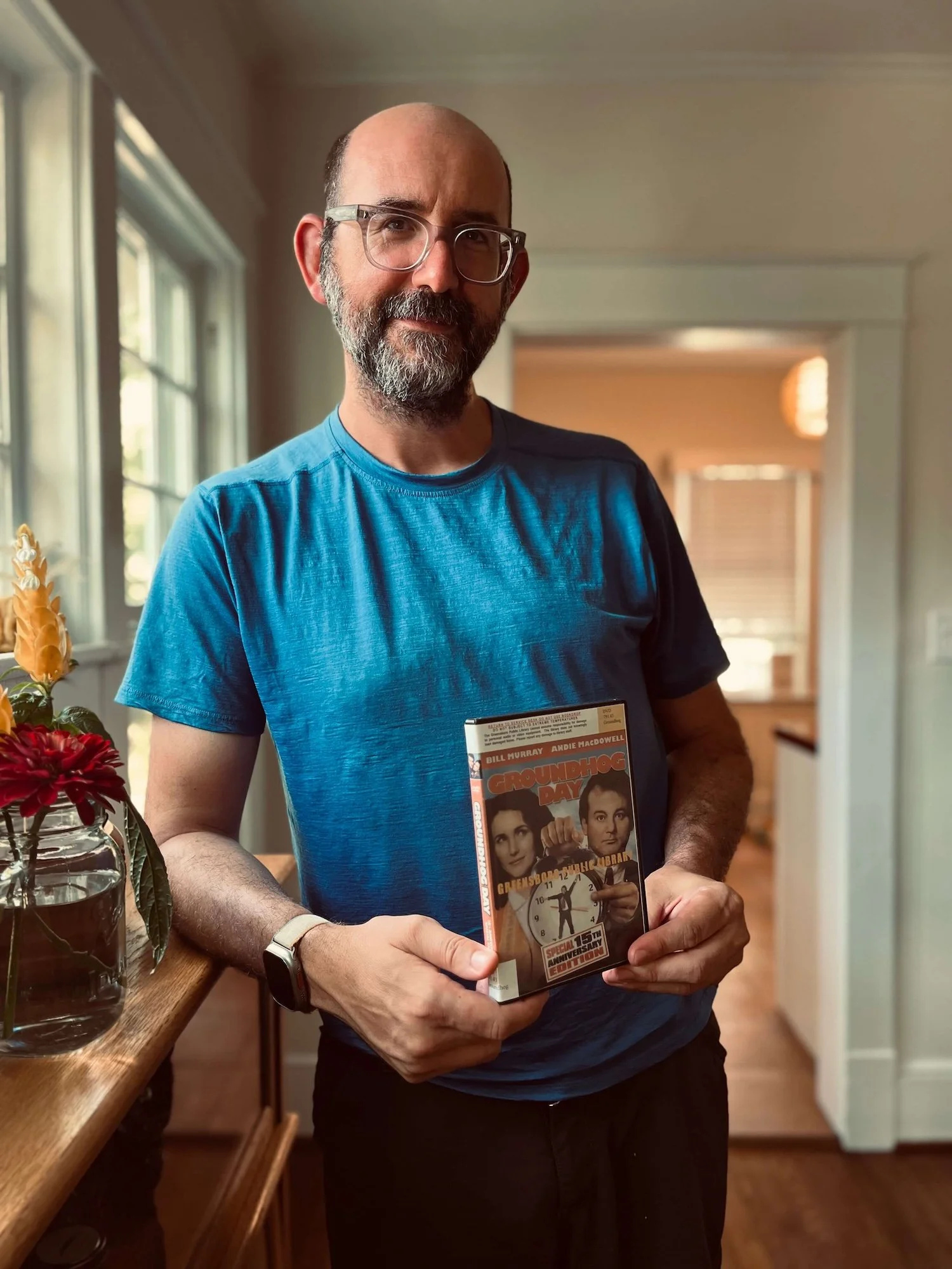 A portrait of a smiling man with glasses and a beard in a blue shirt standing in a bungalow dining room, holding up a Greensboro Public Library DVD copy of the movie Groundhog Day.