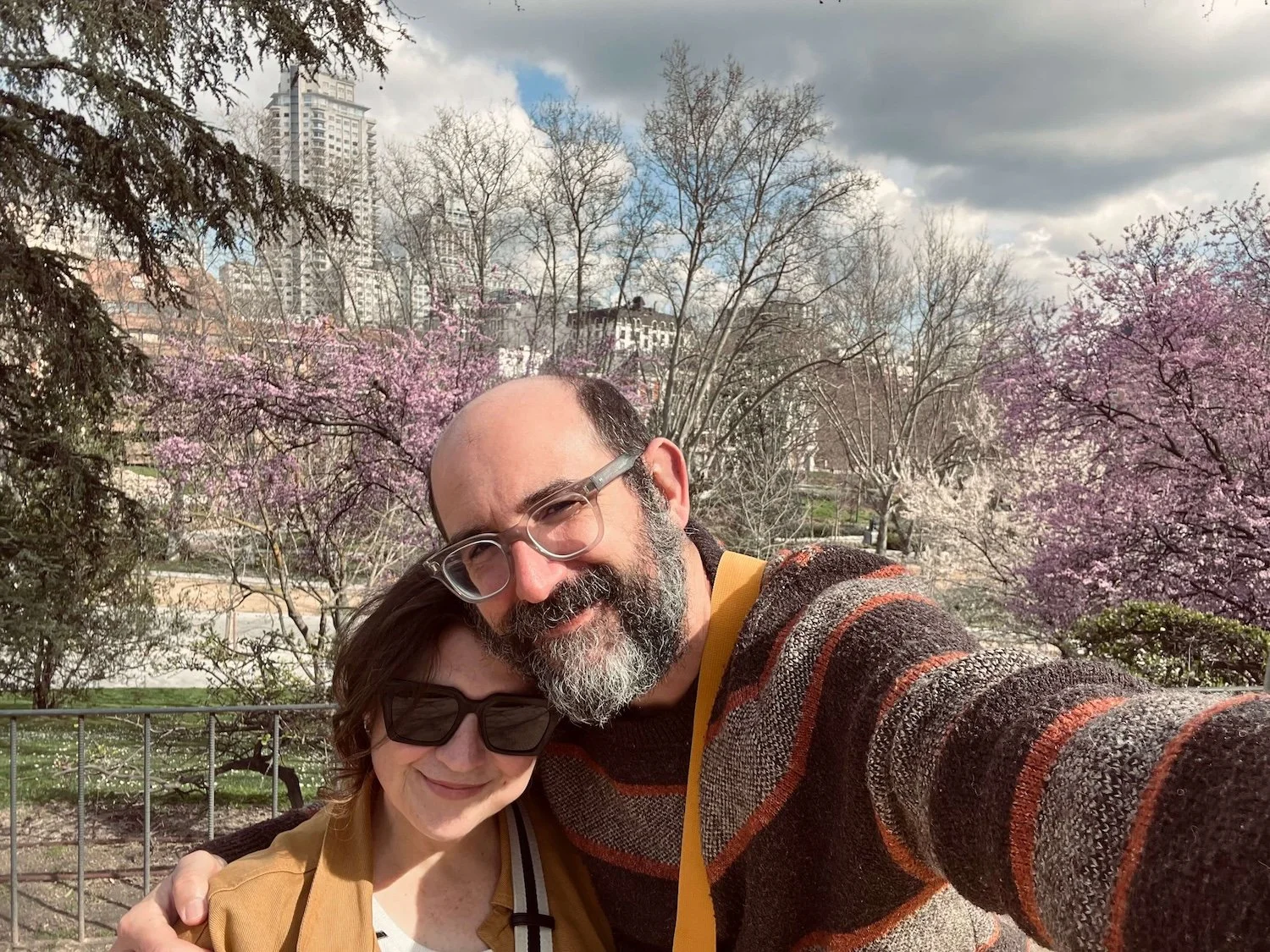 A warm, smiling close-up portrait of the authors of The Dawnatella Edit, a man with glasses and a beard in a striped sweater and a woman in sunglasses and a yellow jacket, with blooming pink trees and buildings in the background.