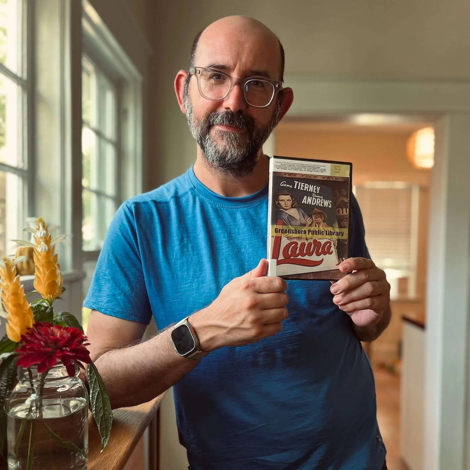 A close-up portrait of a man with glasses and a beard leaning against a barrister bookcase next to a jar of fresh flowers, holding a Greensboro Public Library DVD copy of the classic film noir Laura.