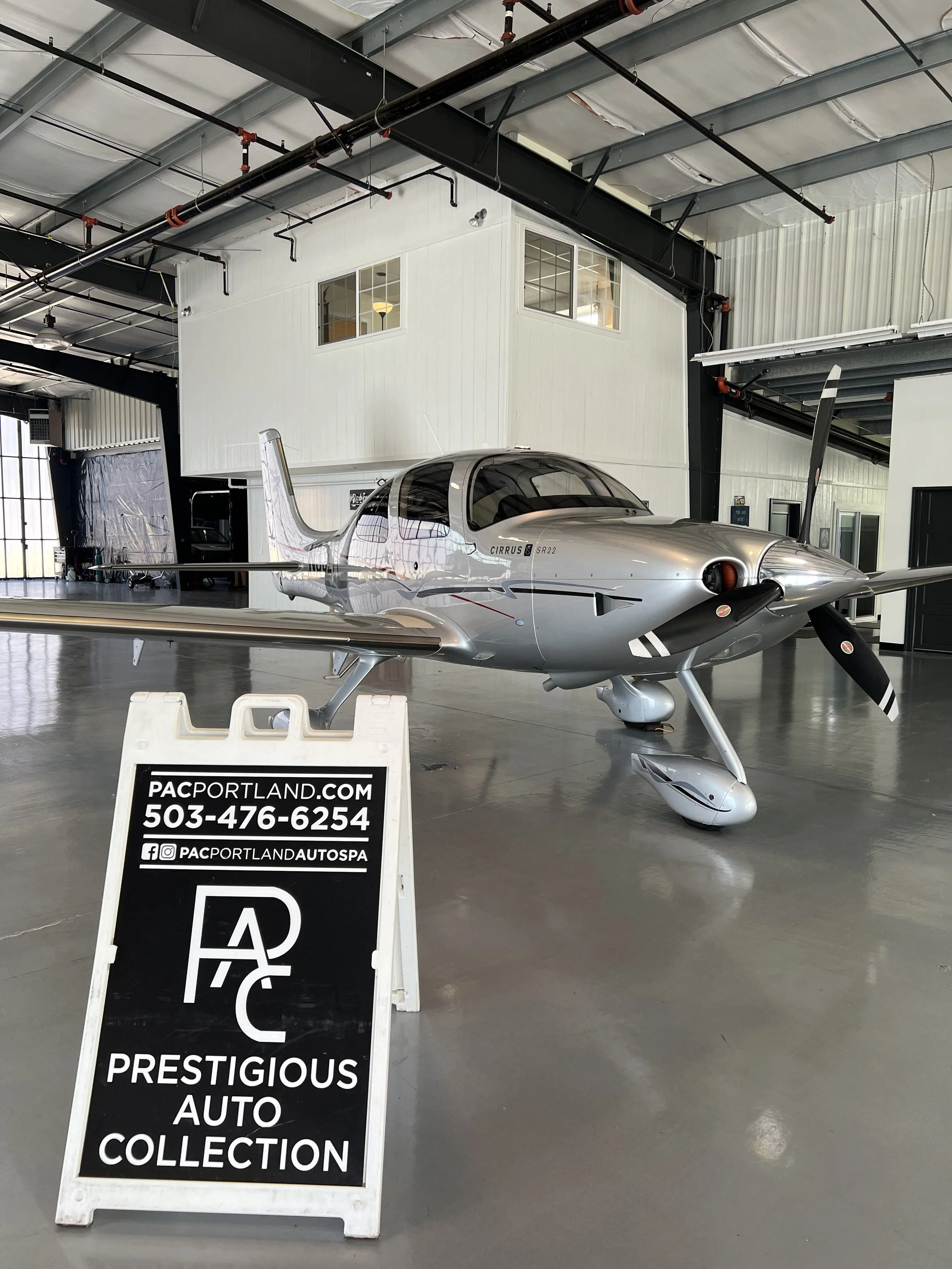 A small silver airplane inside a hangar with a sign in front of it that reads 'Prestigious Auto Collection' and includes contact information.