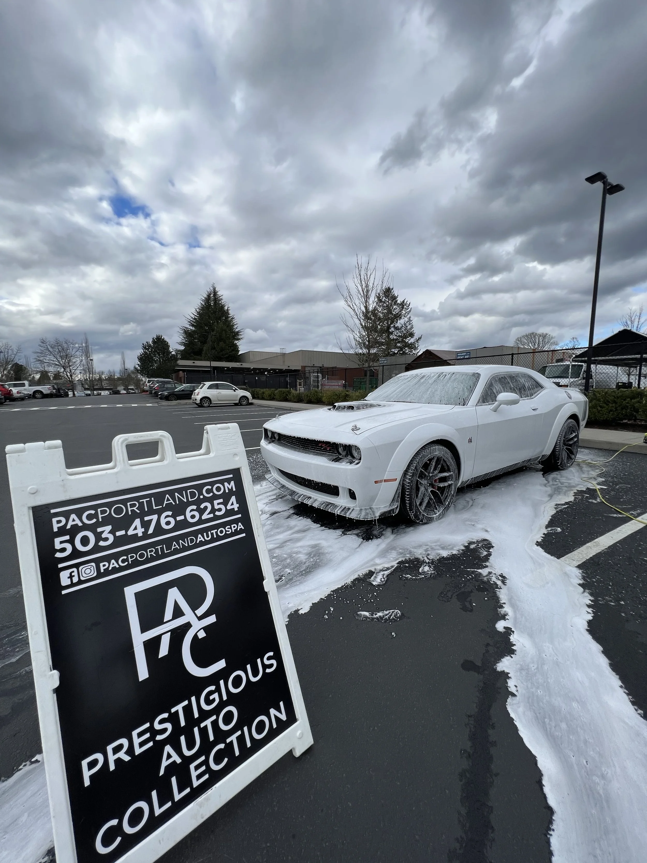 A white Dodge Challenger car is being washed and foamed at a car wash facility. The car is parked on a black asphalt lot with foam and water around it, and a sign for Prestigious Auto Collection with contact information is in the foreground. The background includes other parked cars and a cloudy sky.