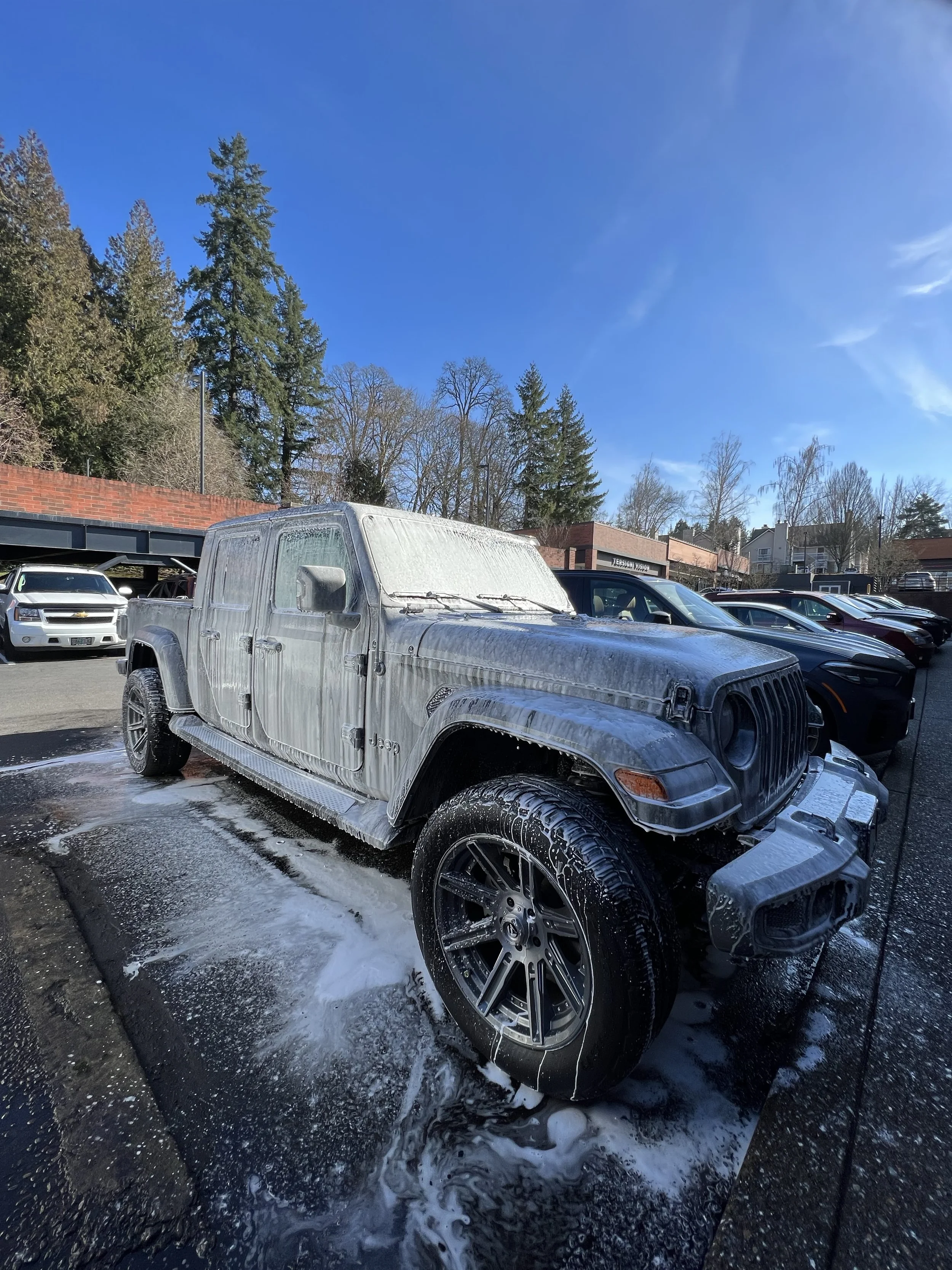 A silver Jeep Gladiator covered in soap and foam during a car wash in a parking lot on a sunny day.