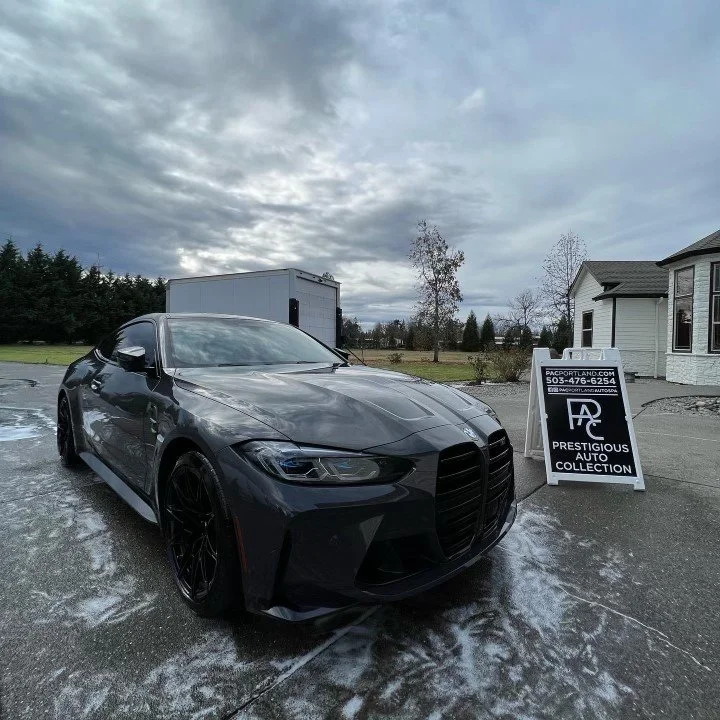 A black sports car parked on a wet driveway under a cloudy sky, with a sign for Prestigious Auto Collection nearby.