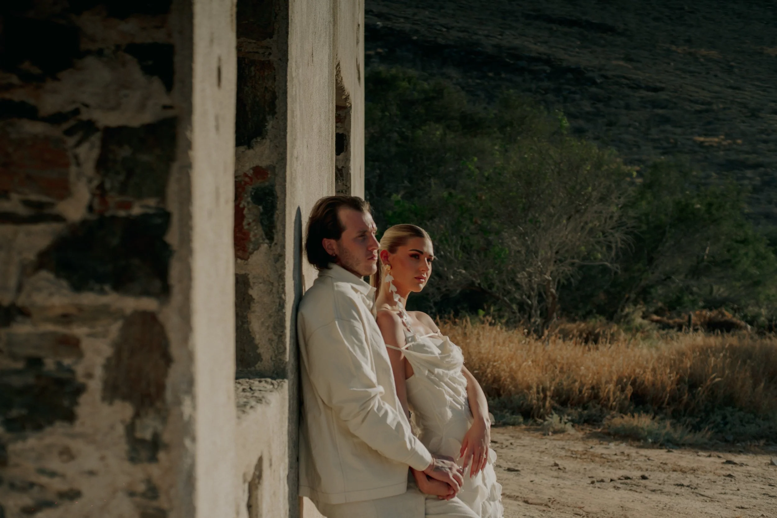 Couple in white clothing leaning against a stone wall in a rustic outdoor setting.