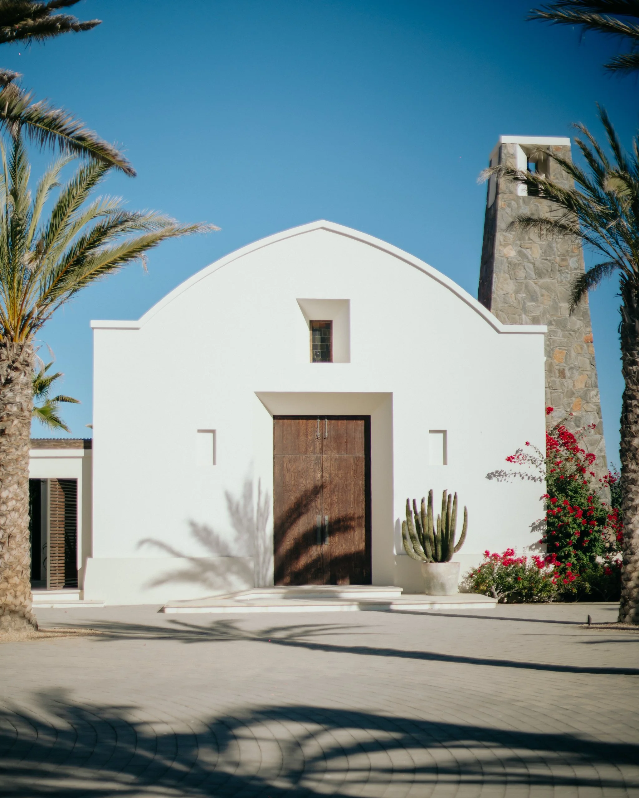 White building with arch, wooden door, stone chimney, palm trees, and cactus in planter, under clear blue sky.