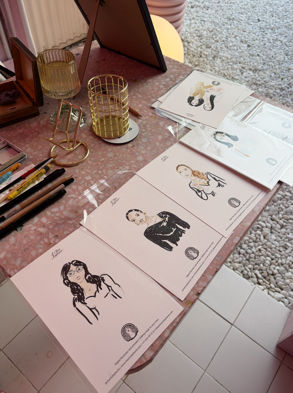 Een overzicht van getekende portretten van verschillende vrouwen, uitgestald op een roze tafel met tekenmaterialen en accessoires.