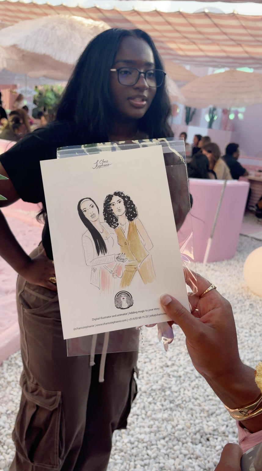 Een vrouw met lang zwart haar en brillen houdt een illustratie van twee vrouwen. De afbeelding wordt vastgehouden door iemand die rechtsonder in de foto met een gouden ring en armband te zien is. De setting is een kleurrijke, gezellige binnenruimte met parasols en mensen op de achtergrond.