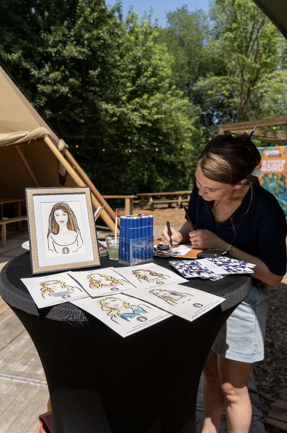 Vrouw maakt karikaturen op een outdoor evenement, met tekeningen van haar kunstwerk op tafel en een portret van een vrouw in een lijst.